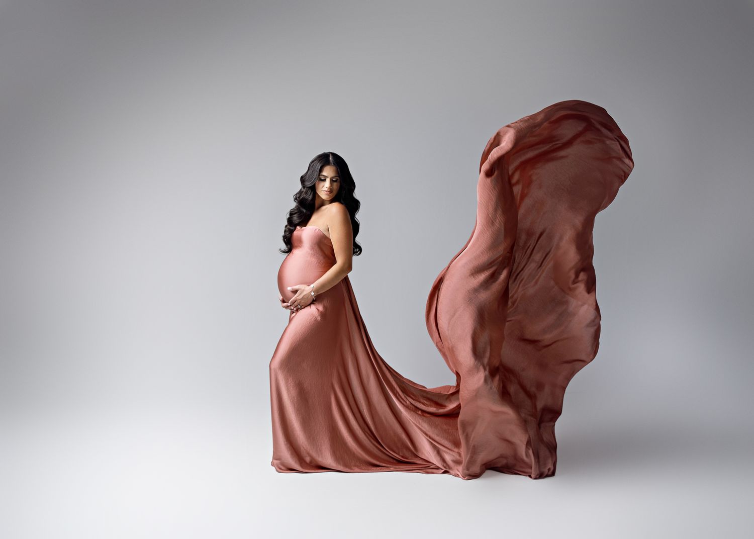 Artistic maternity photo showing a person in flowing dusty rose gown against gray backdrop with fabric billowing dramatically.