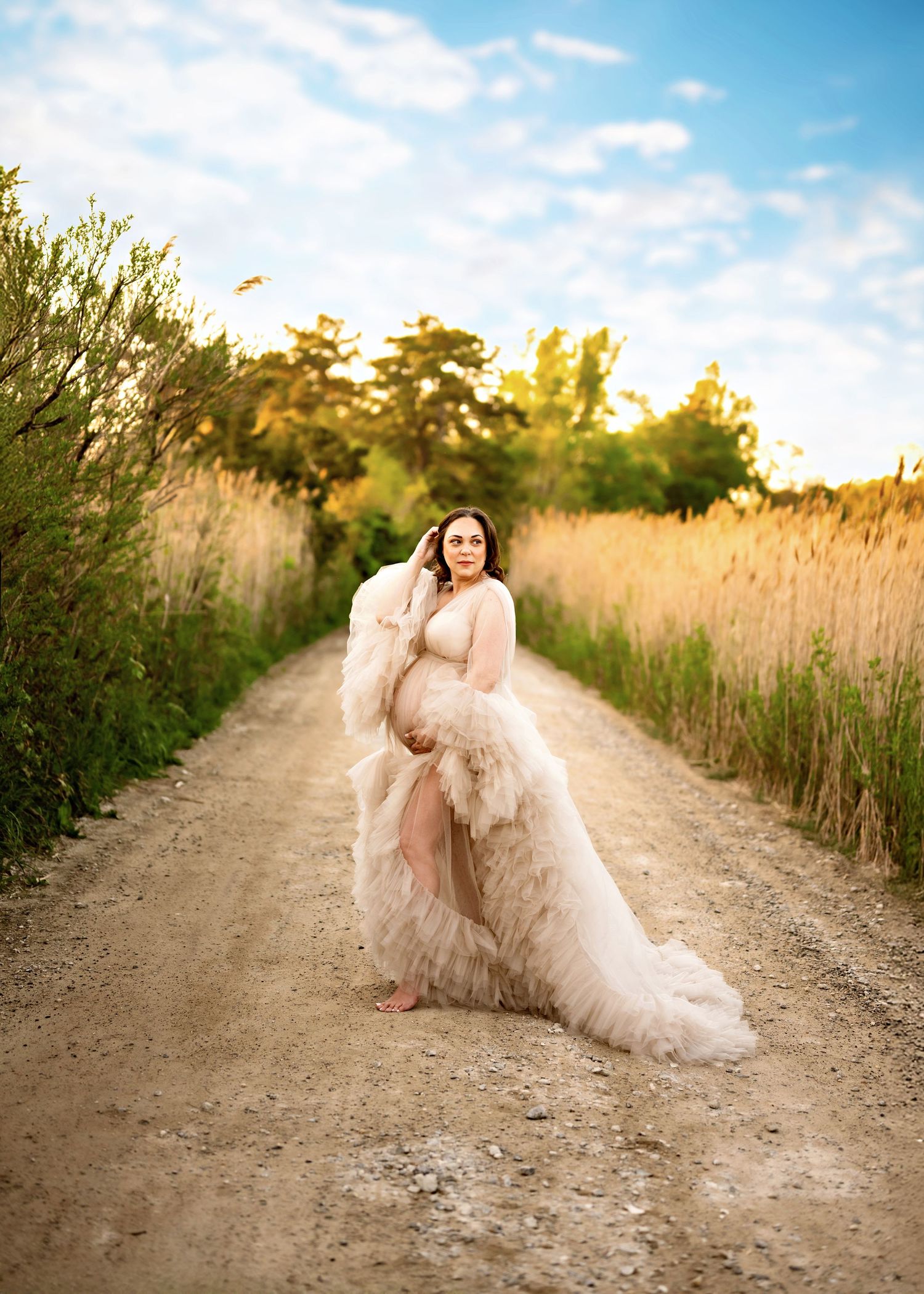 A flowing cream-colored dress billows dramatically on a dirt path between tall grasses during golden hour.
