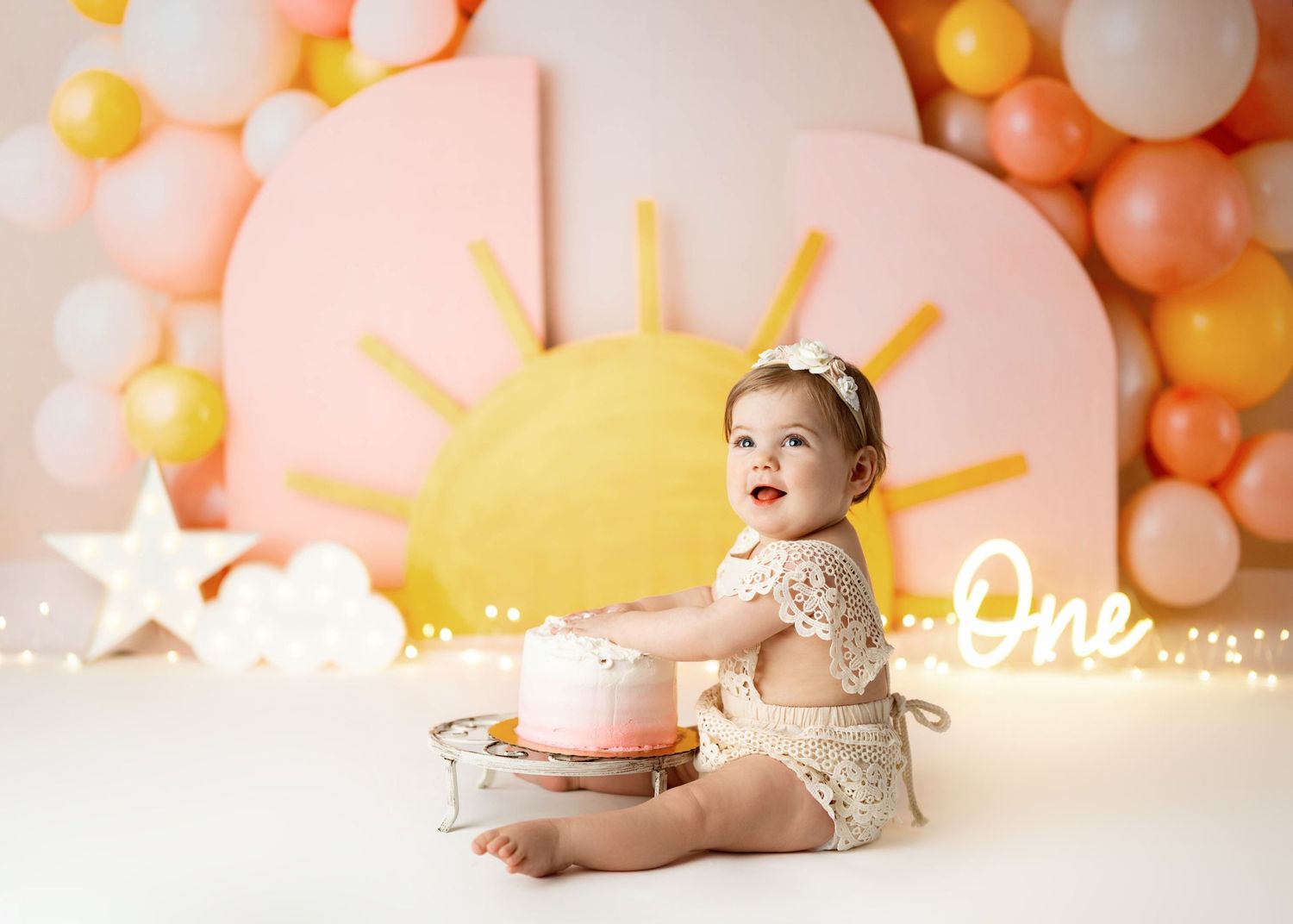 Whimsical first birthday cake smash setup with pink and yellow decor, balloons, and star decorations against pastel backdrop.
