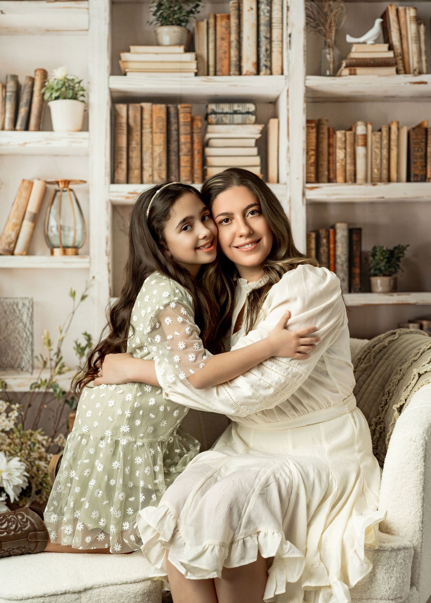 Two people in white and polka dot dresses embrace while sitting on a couch in front of built-in bookshelves.