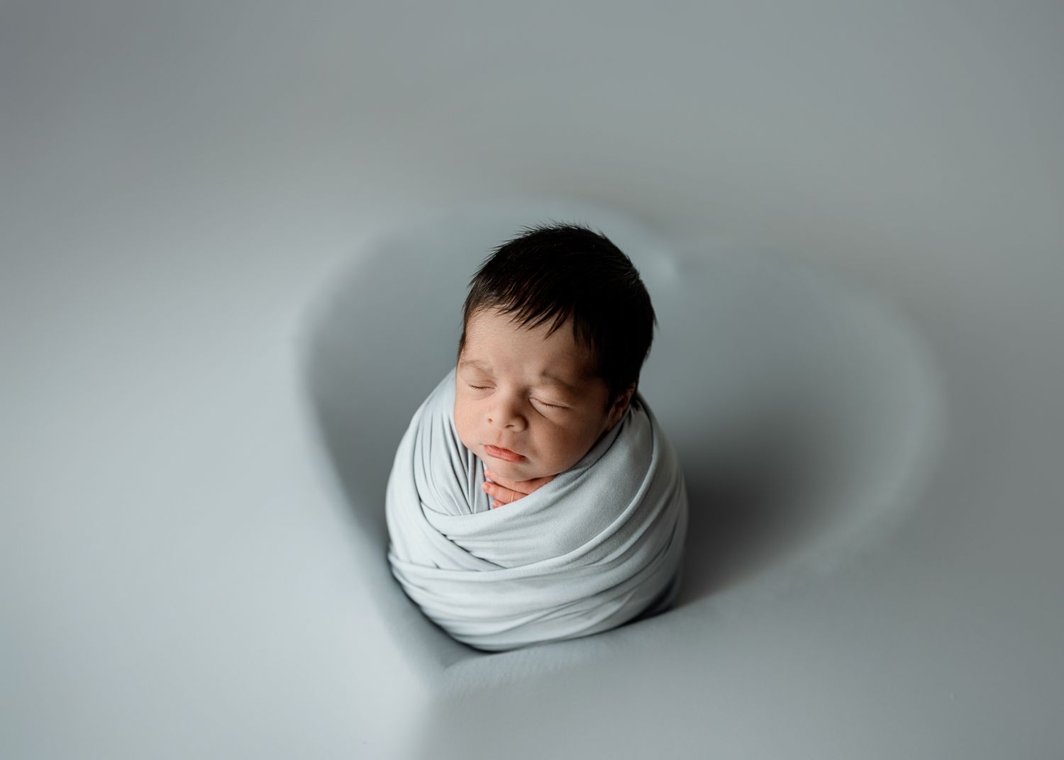 Newborn baby peacefully sleeping while wrapped in soft white swaddle against neutral grey background.