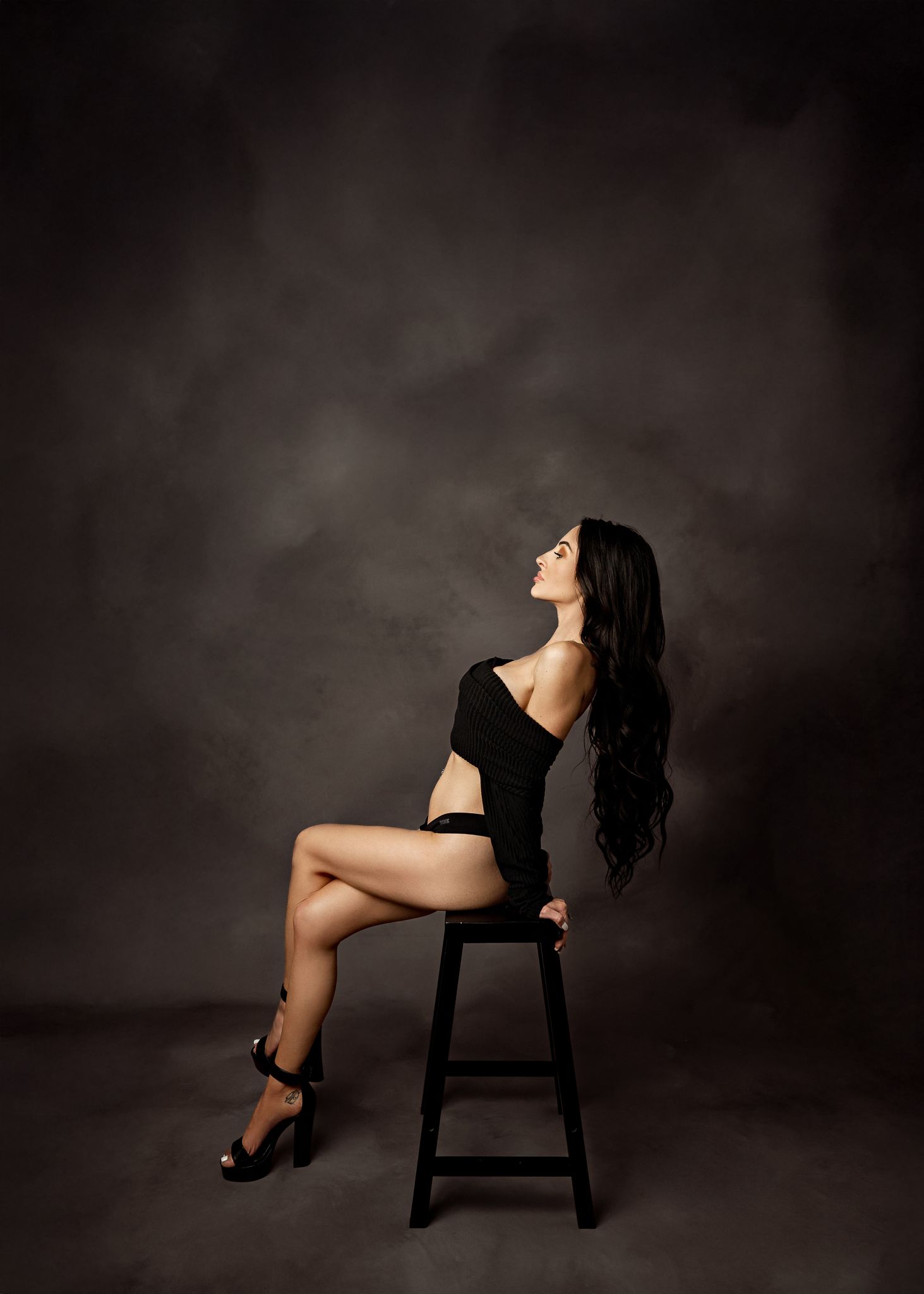Model with long dark hair poses elegantly on wooden stool against moody dark background in studio setting.