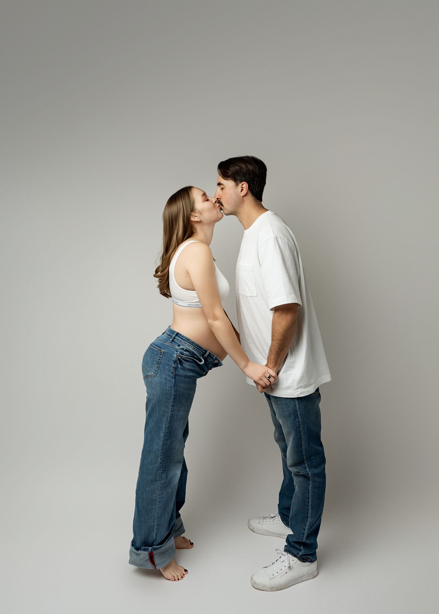 A couple in matching white shirts and jeans share a tender kiss against a gray backdrop in a minimalist studio setting.
