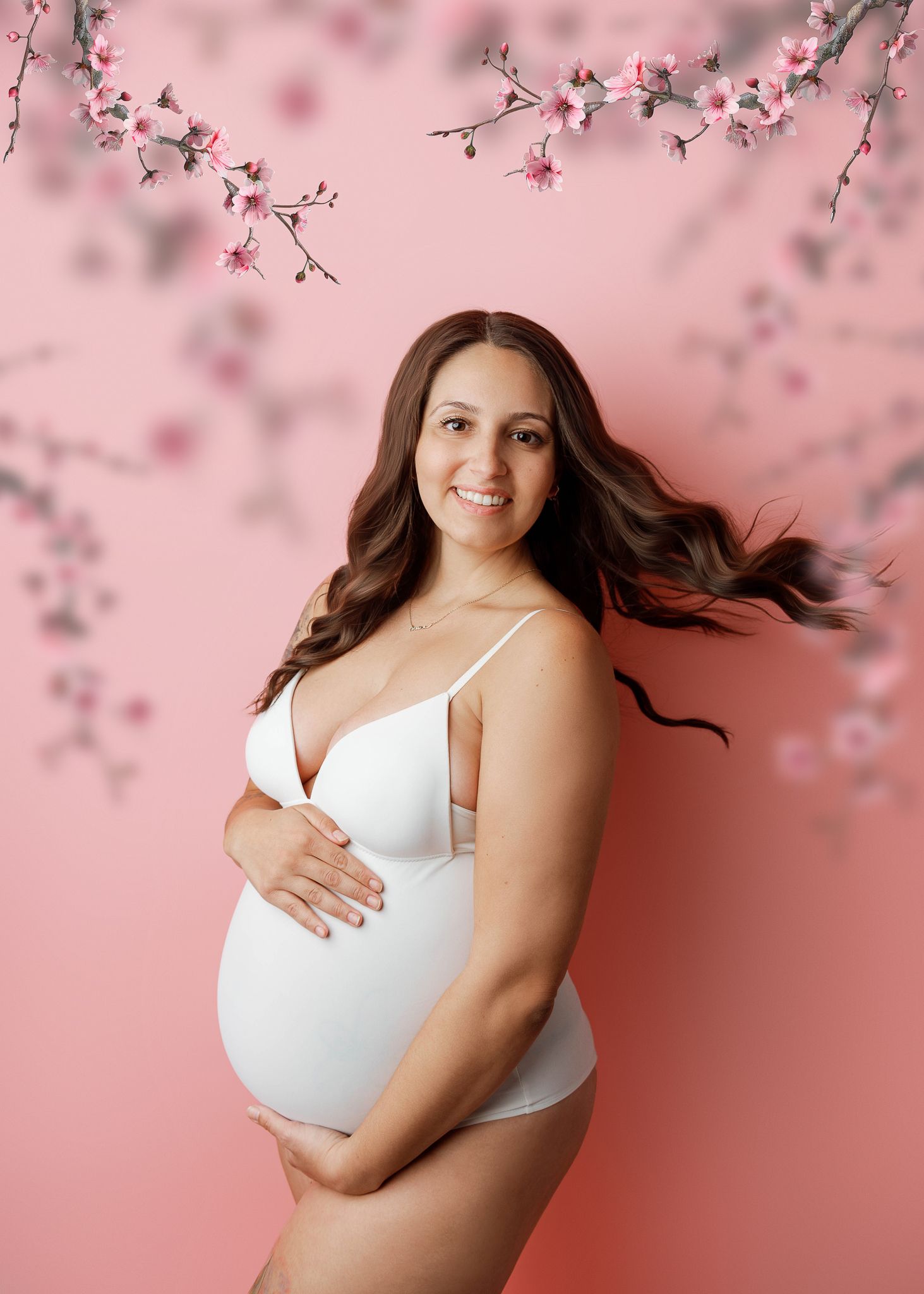 Maternity photo with cherry blossoms against pink background showing expectant mother in white lingerie.