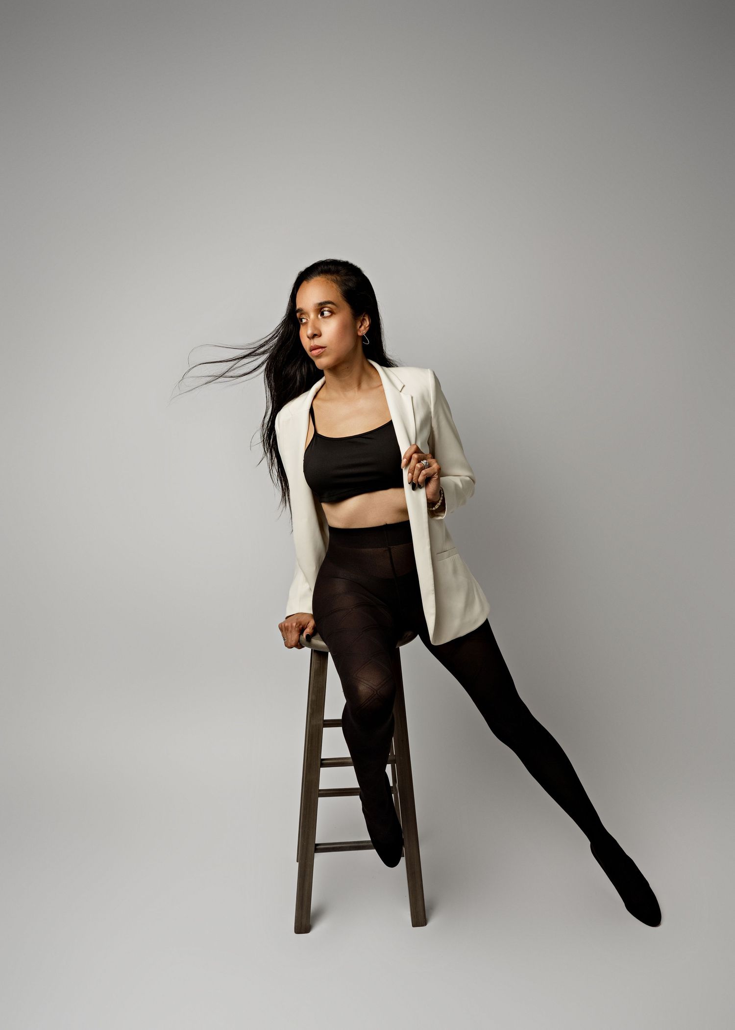 Dancer in black athletic wear and white blazer poses dynamically on wooden stool against neutral background.