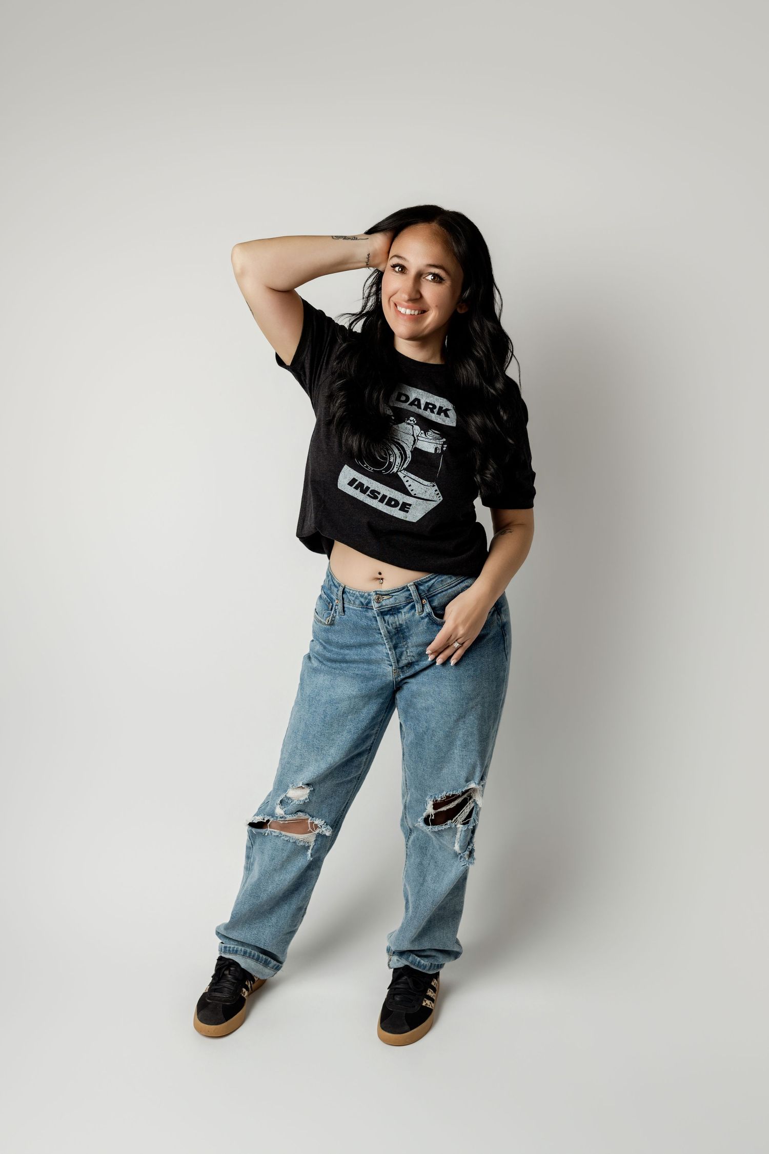 Woman in black graphic tee and ripped jeans posing confidently in a studio setting with a white background.