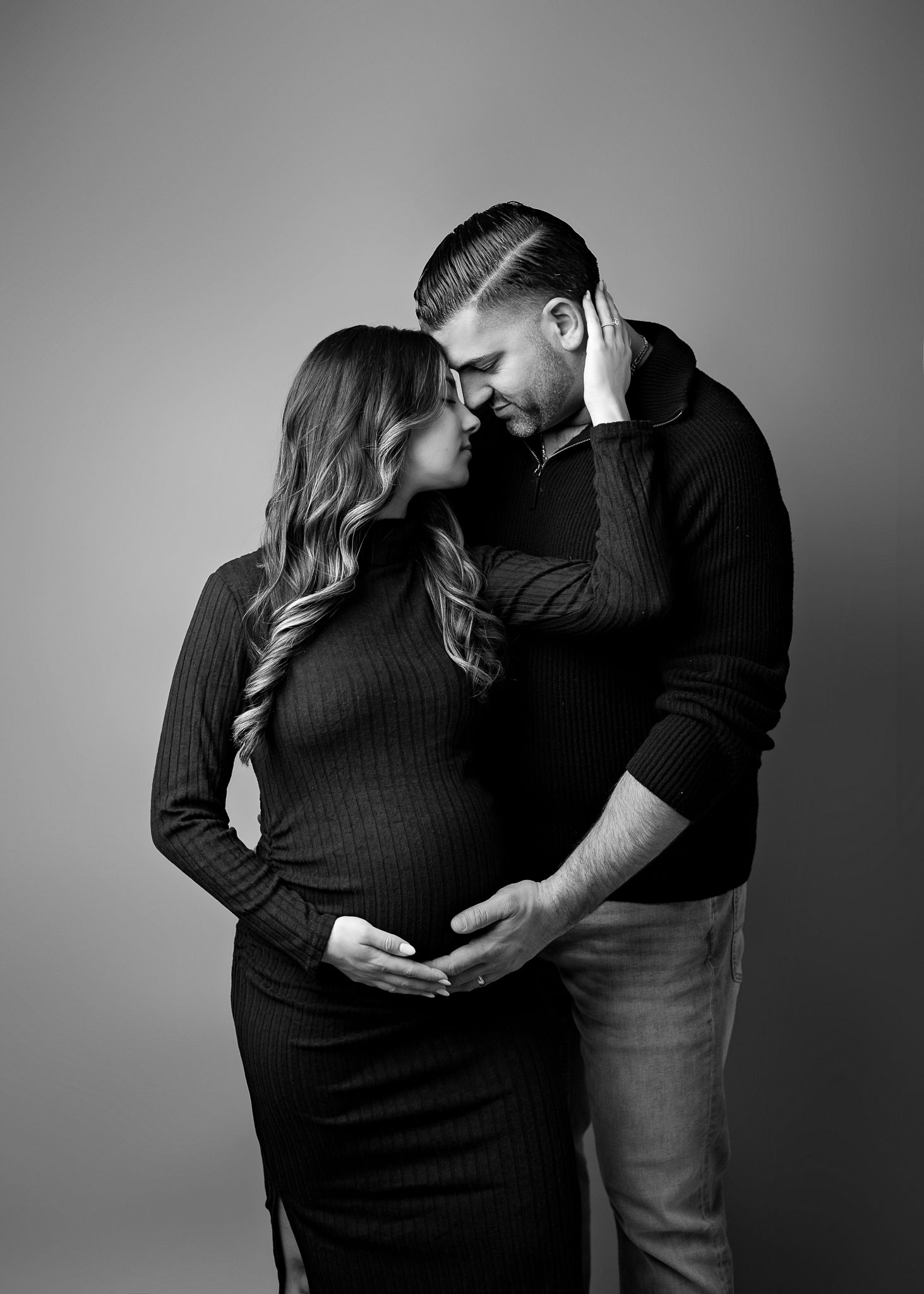 Intimate black and white maternity photo shows couple in dark clothing embracing tenderly against neutral background.