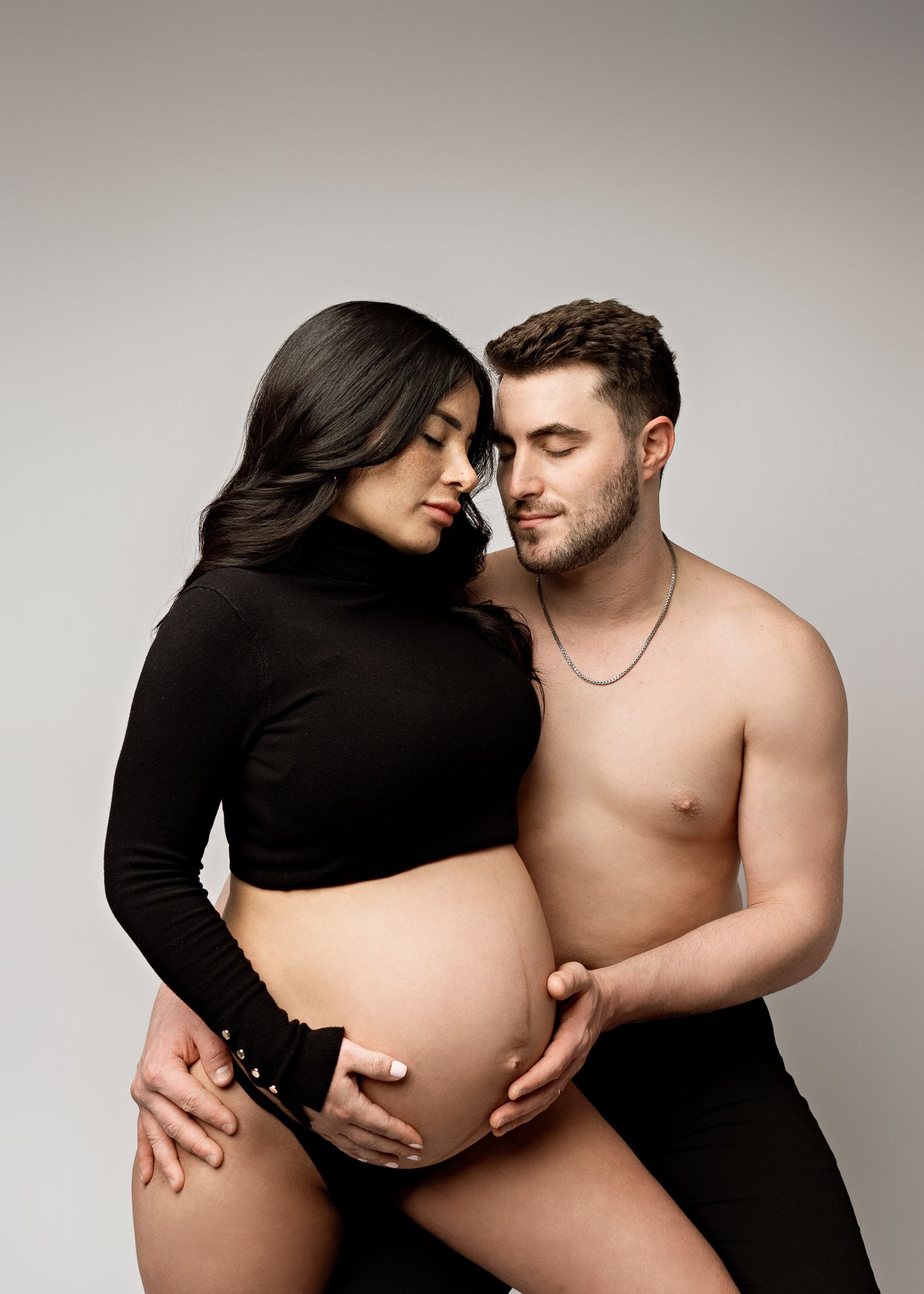 An intimate maternity photo shows a couple embracing, with her in a black turtleneck and him shirtless against a neutral backdrop.