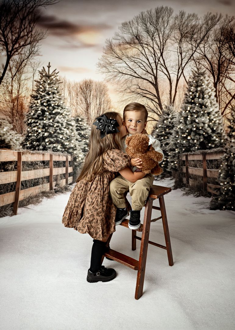 Two children in a snowy Christmas tree farm setting, girl in brown dress hugging boy on stool holding teddy bear.