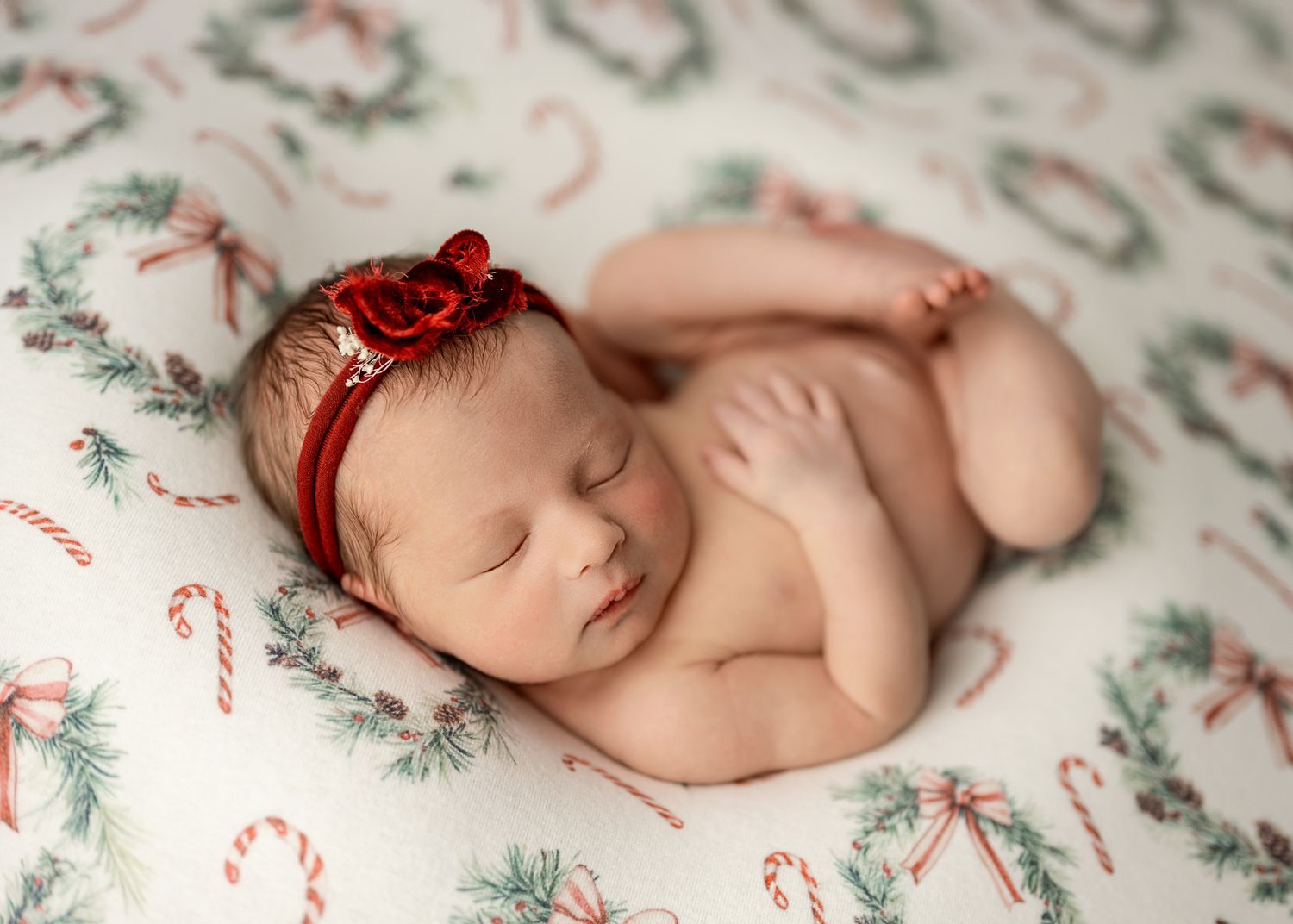 Sleeping newborn on festive Christmas-themed blanket with candy cane and pine branch pattern.