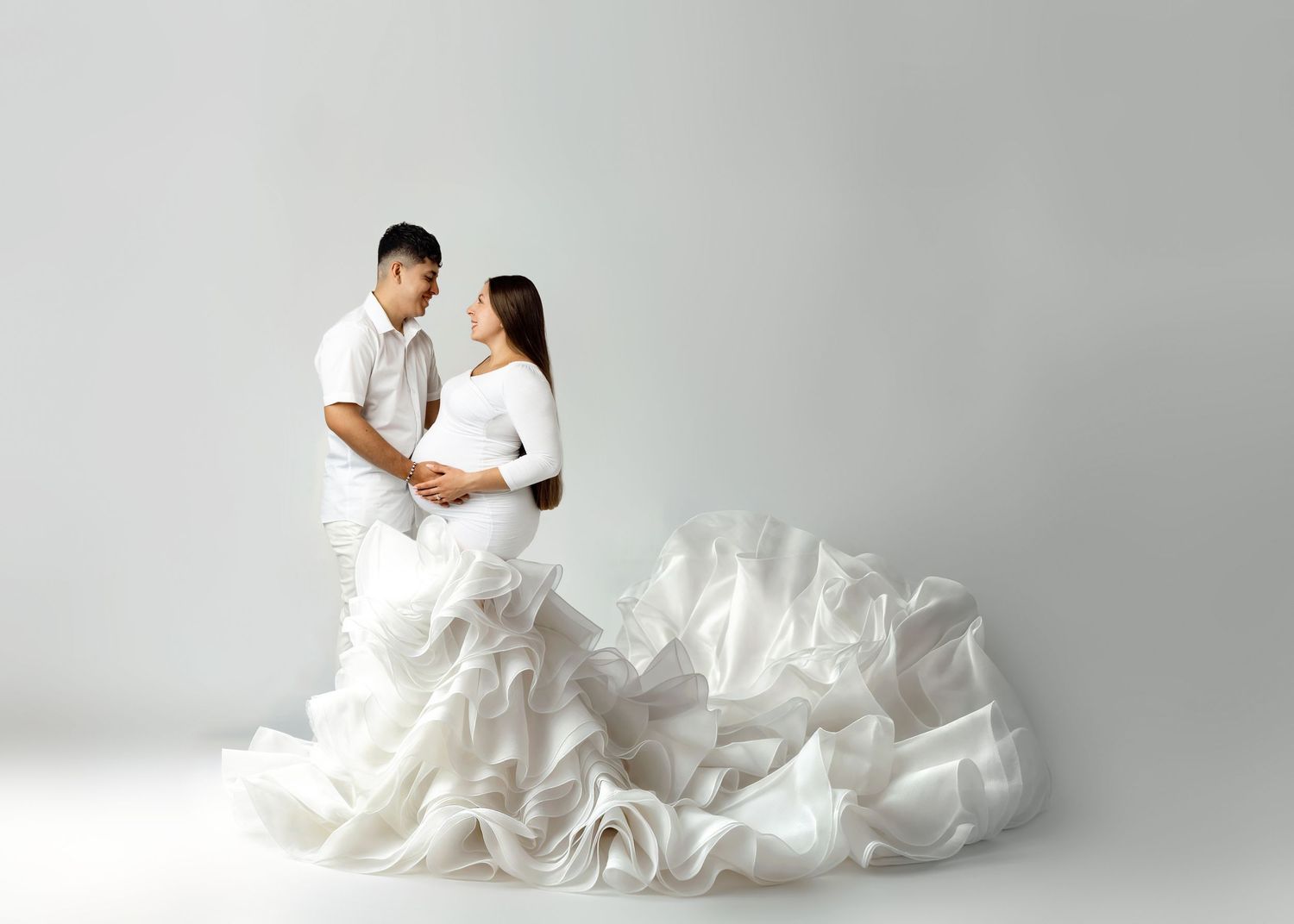 Two figures in white embrace against a light background with flowing white fabric billowing around them in an artistic portrait.