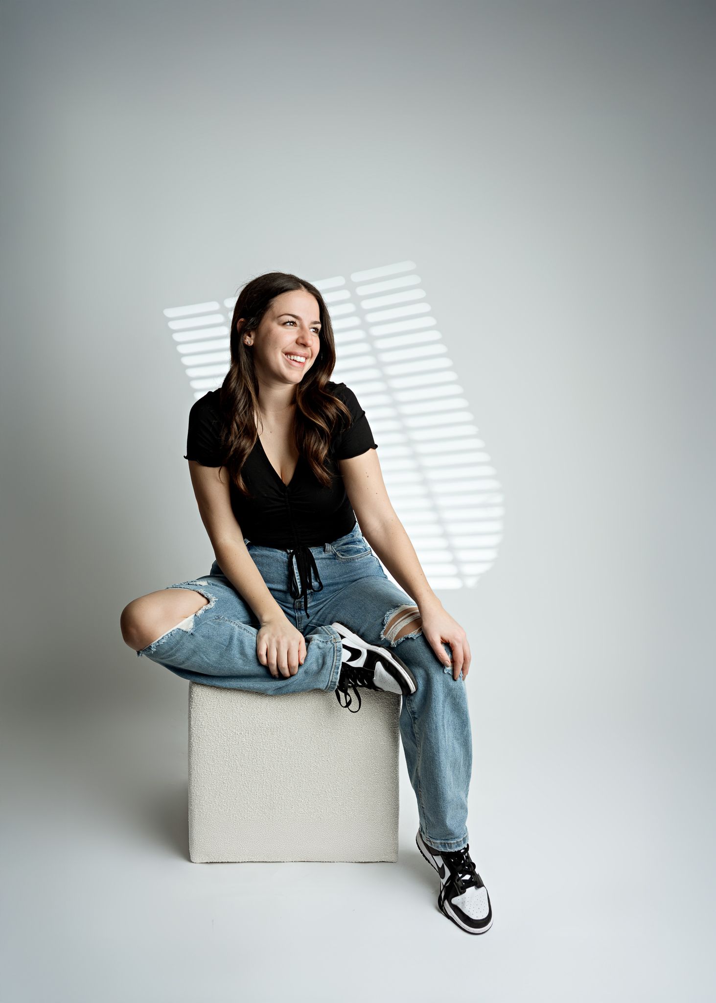 Person in casual attire of black top and distressed jeans sits relaxed on white cube against minimalist gray background.