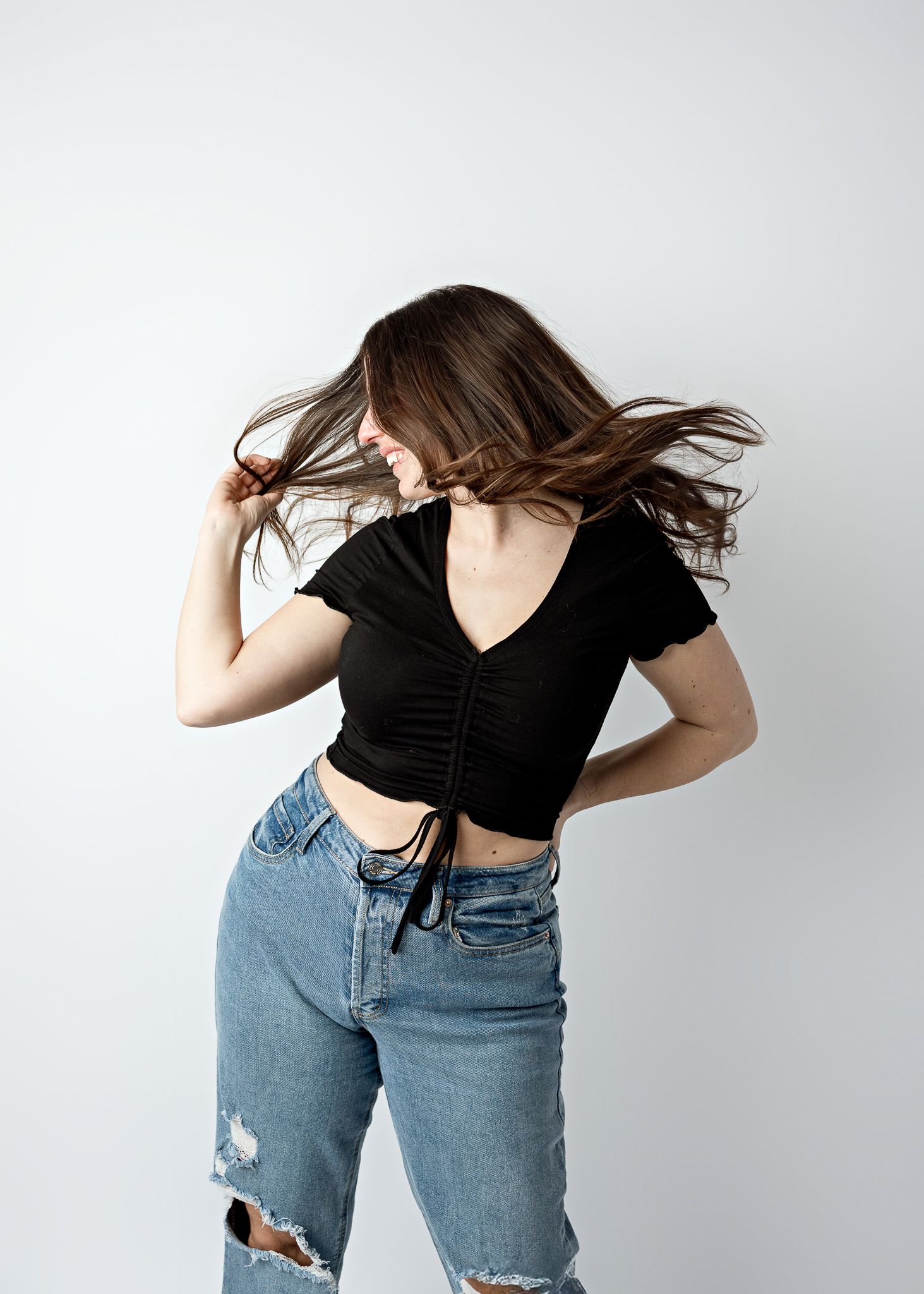Young woman in black crop top and distressed jeans flips hair against white background.