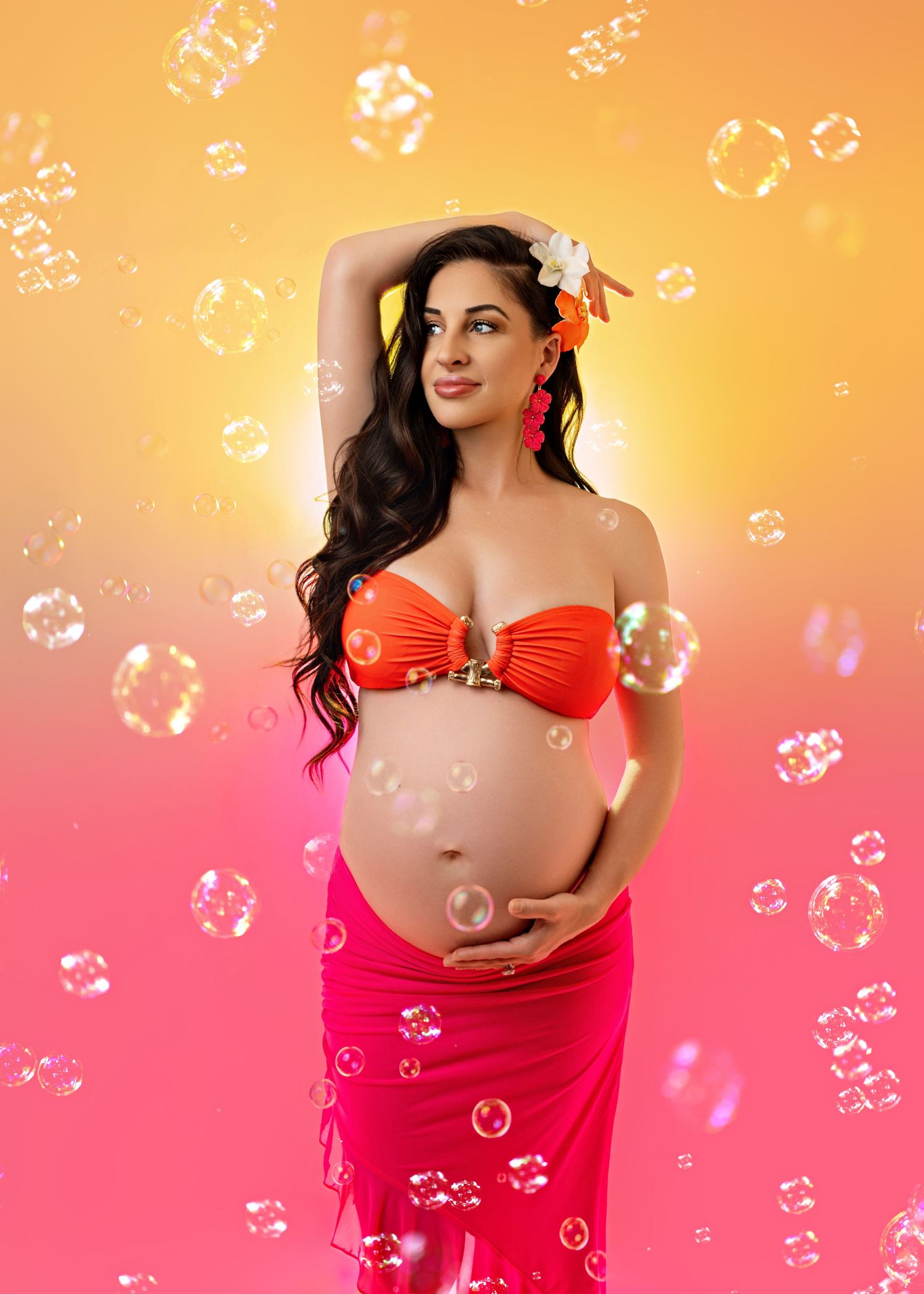 Pregnant woman in orange bikini top and pink sarong poses gracefully with bubbles on yellow-pink gradient background.