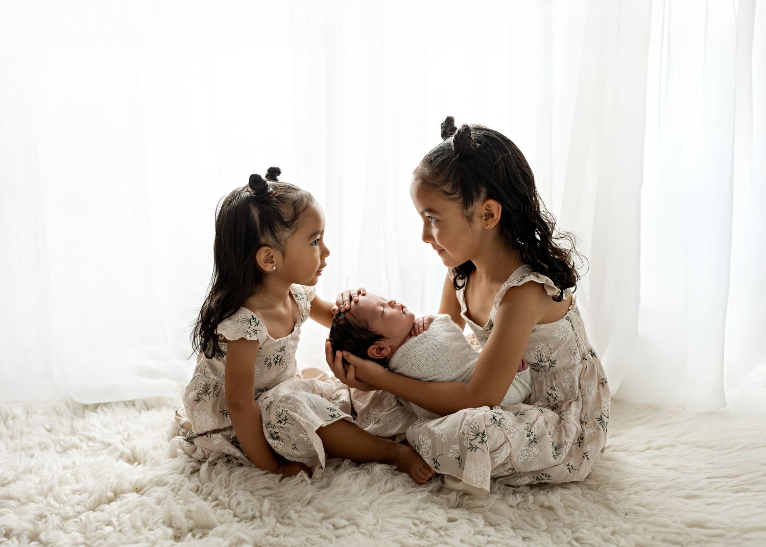 Two children in white lace dresses play together on a light colored floor in a bright studio setting.