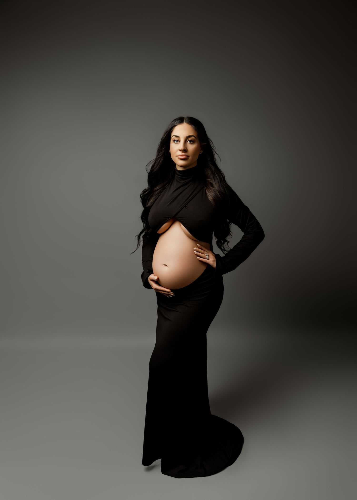 Pregnant woman in black cutout gown cradling baby bump in professional maternity studio portrait.