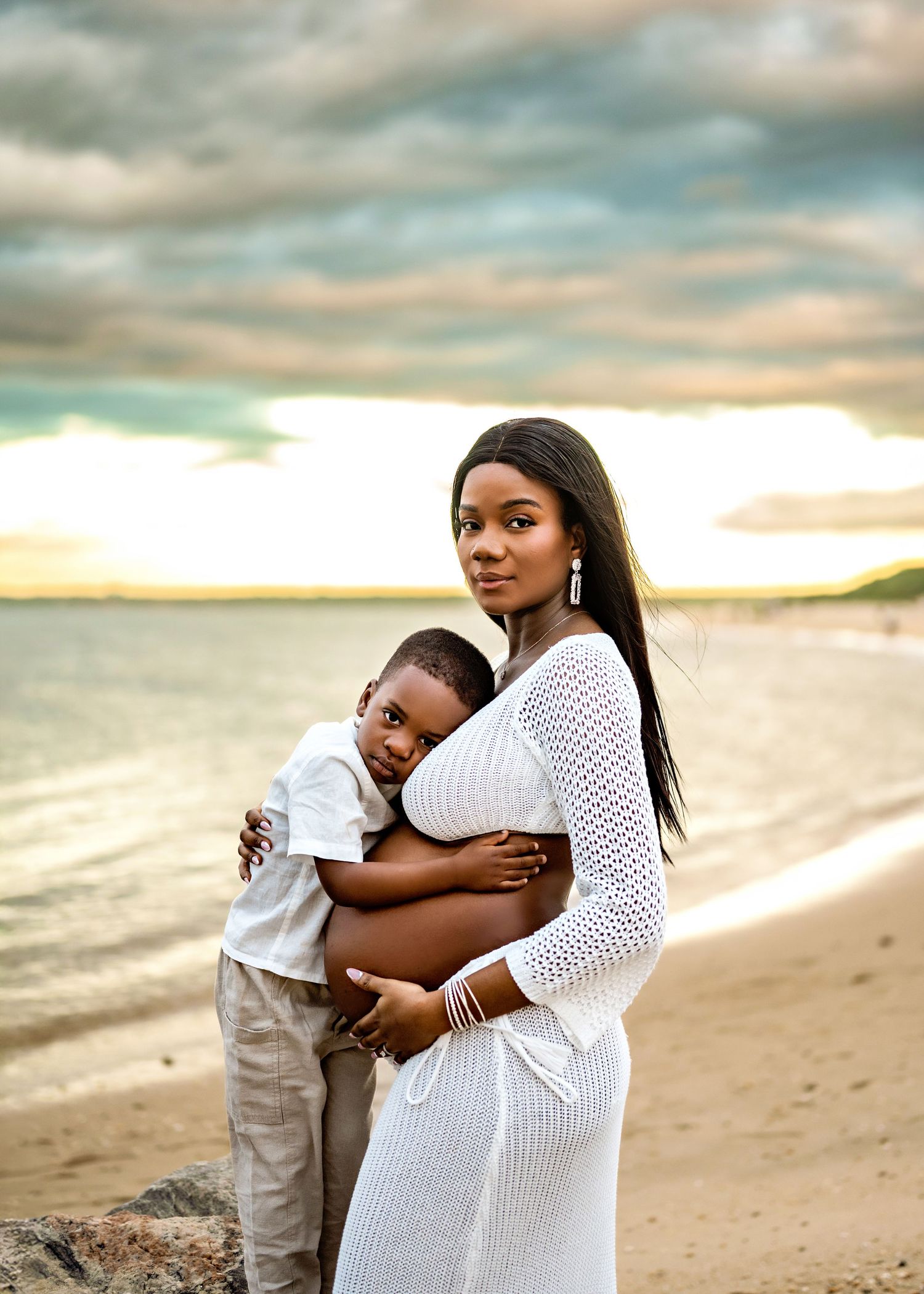 A tender beach portrait at sunset shows maternal love with white knit dress and beige pants against a golden sky.