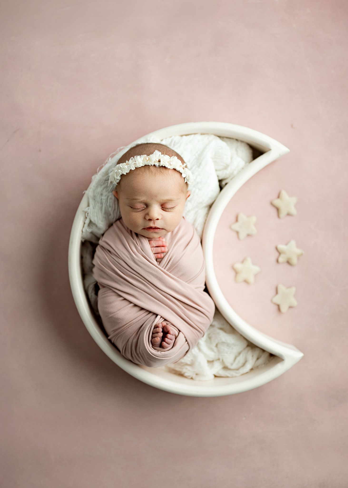 A sleeping newborn wrapped in a pink swaddle rests in a decorative white crescent moon prop with star accents on pink backdrop.