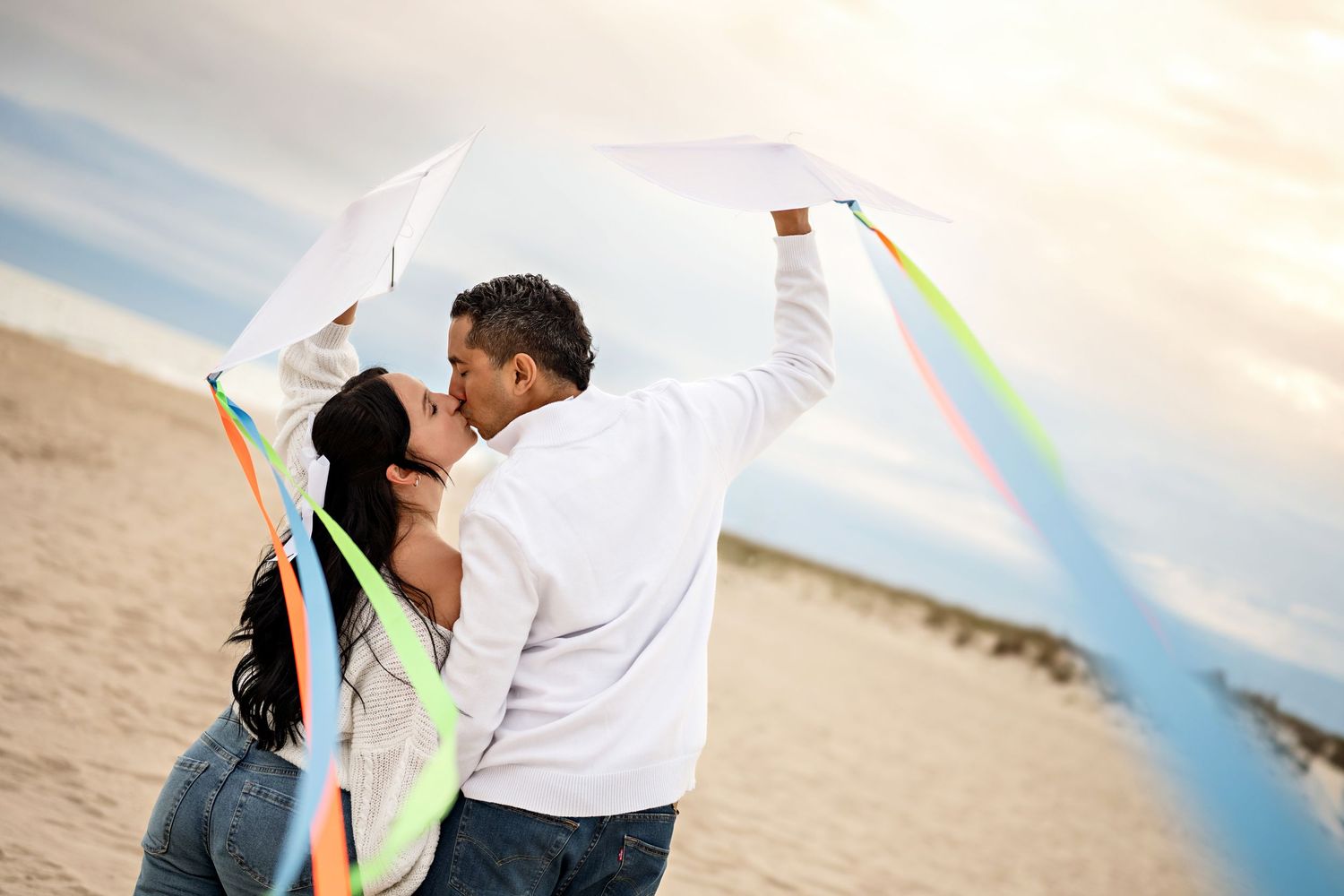 Couple kissing on a sandy beach while holding a colorful kite with ribbons under a cloudy sky.