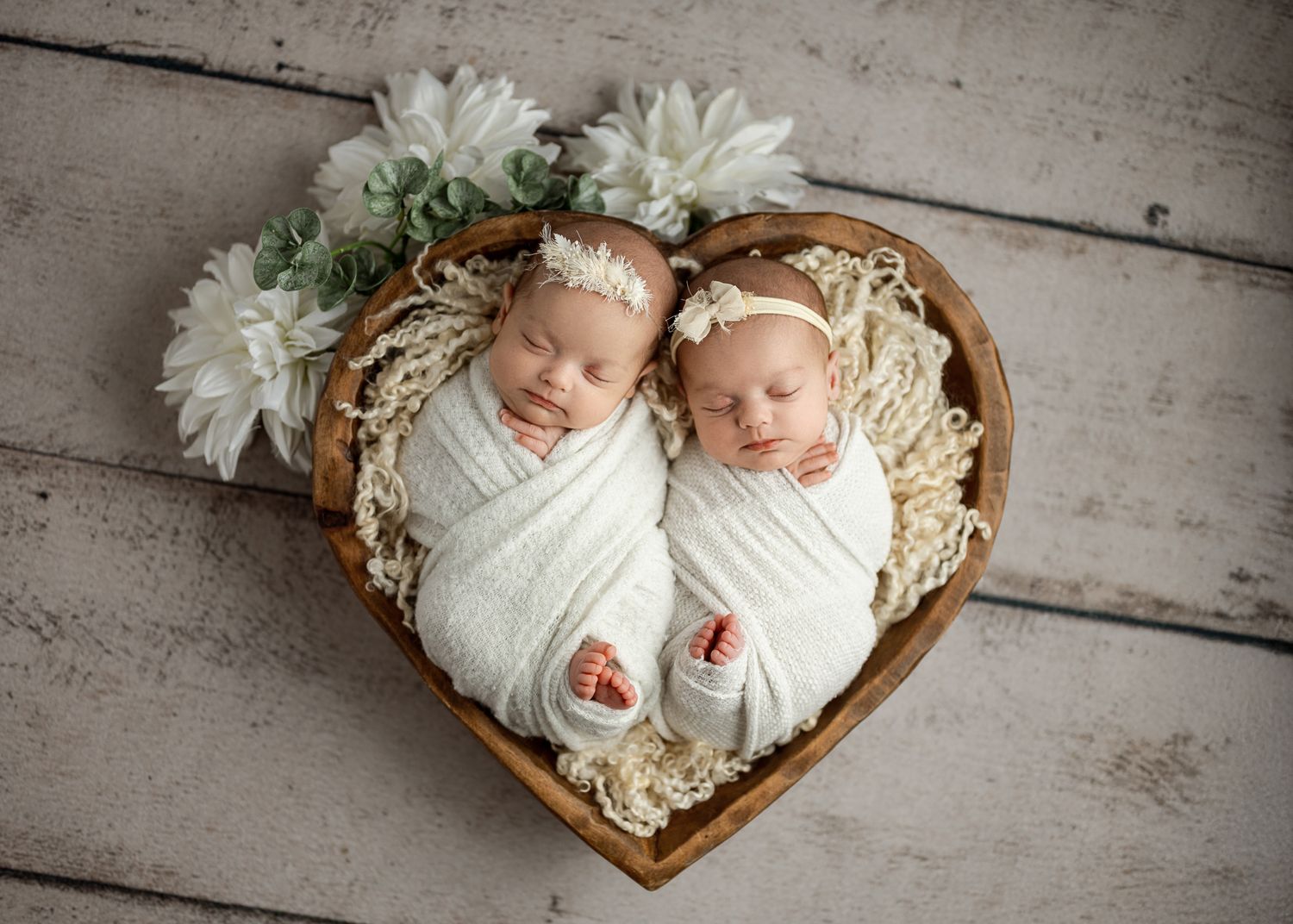 Two swaddled babies sleeping in a heart-shaped wooden basket with white flowers on a rustic wooden floor.