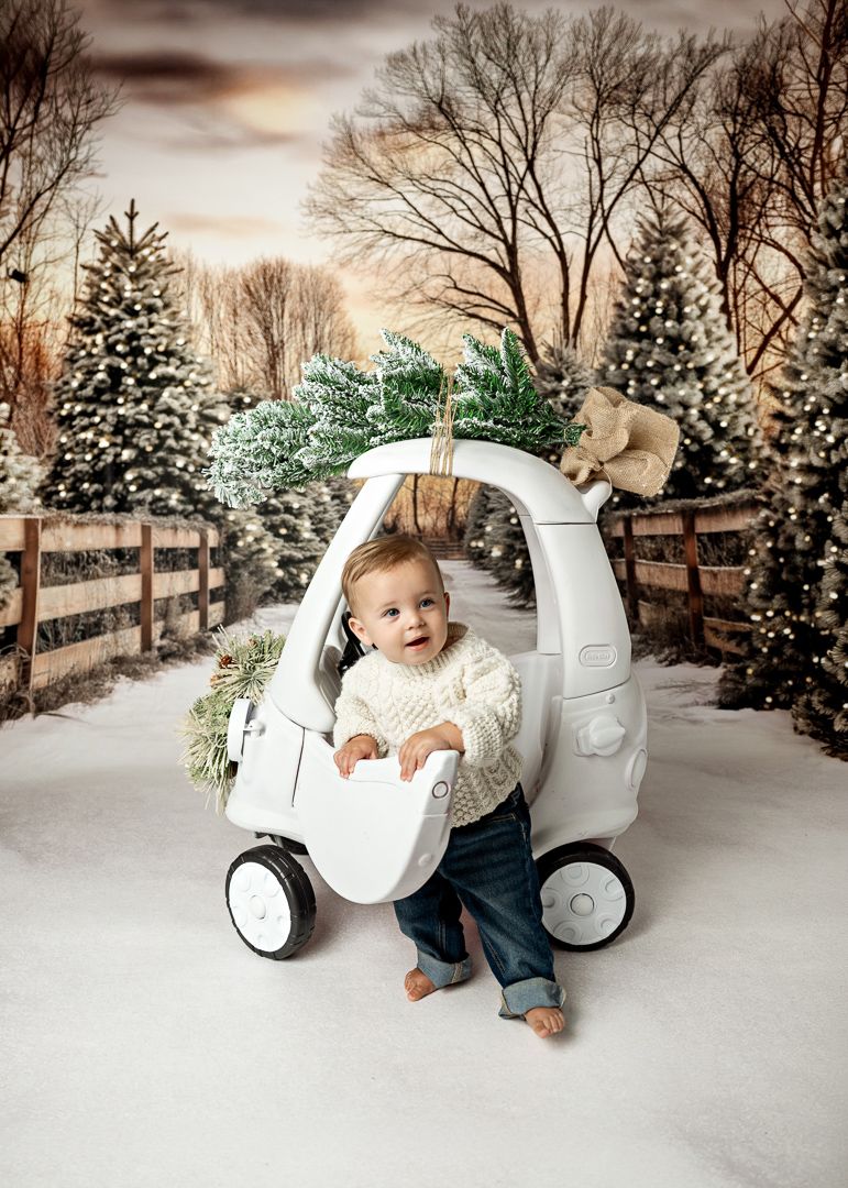 Baby in white toy car with Christmas trees and snow in a festive winter holiday photo session.