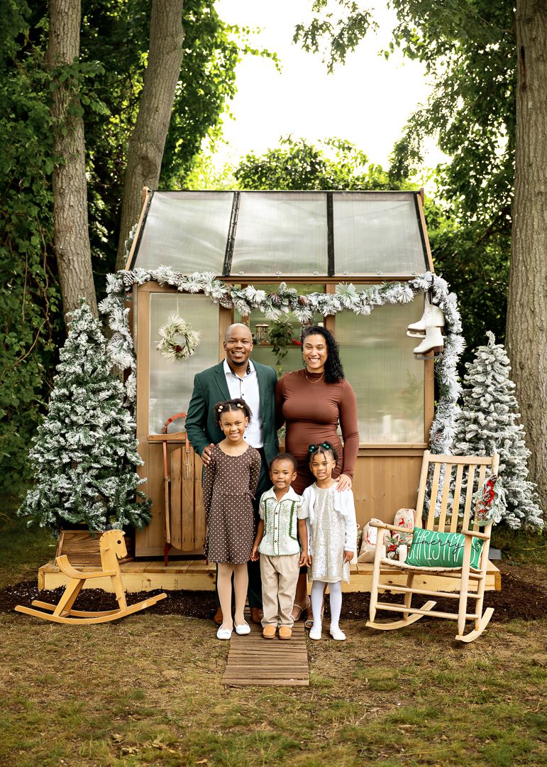 Family of five poses in front of decorated greenhouse with Christmas trees and holiday garland in backyard.