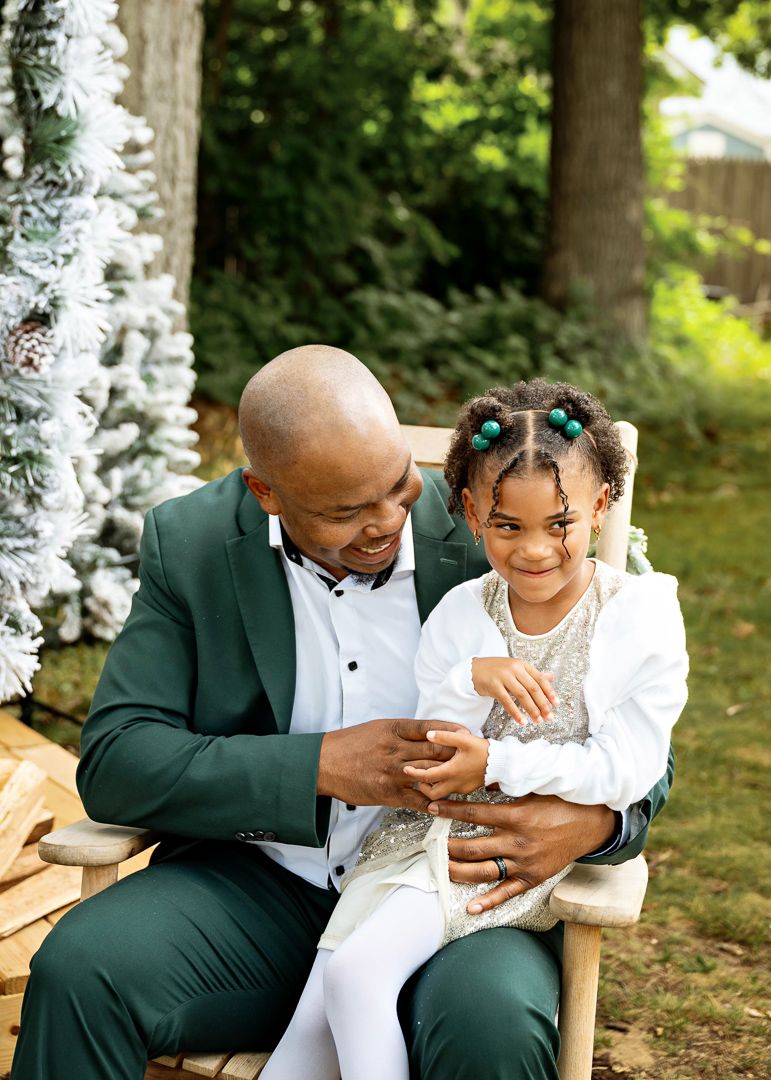 A bald man in a green tuxedo sits outdoors holding a young girl with colorful hair accessories in a white outfit.