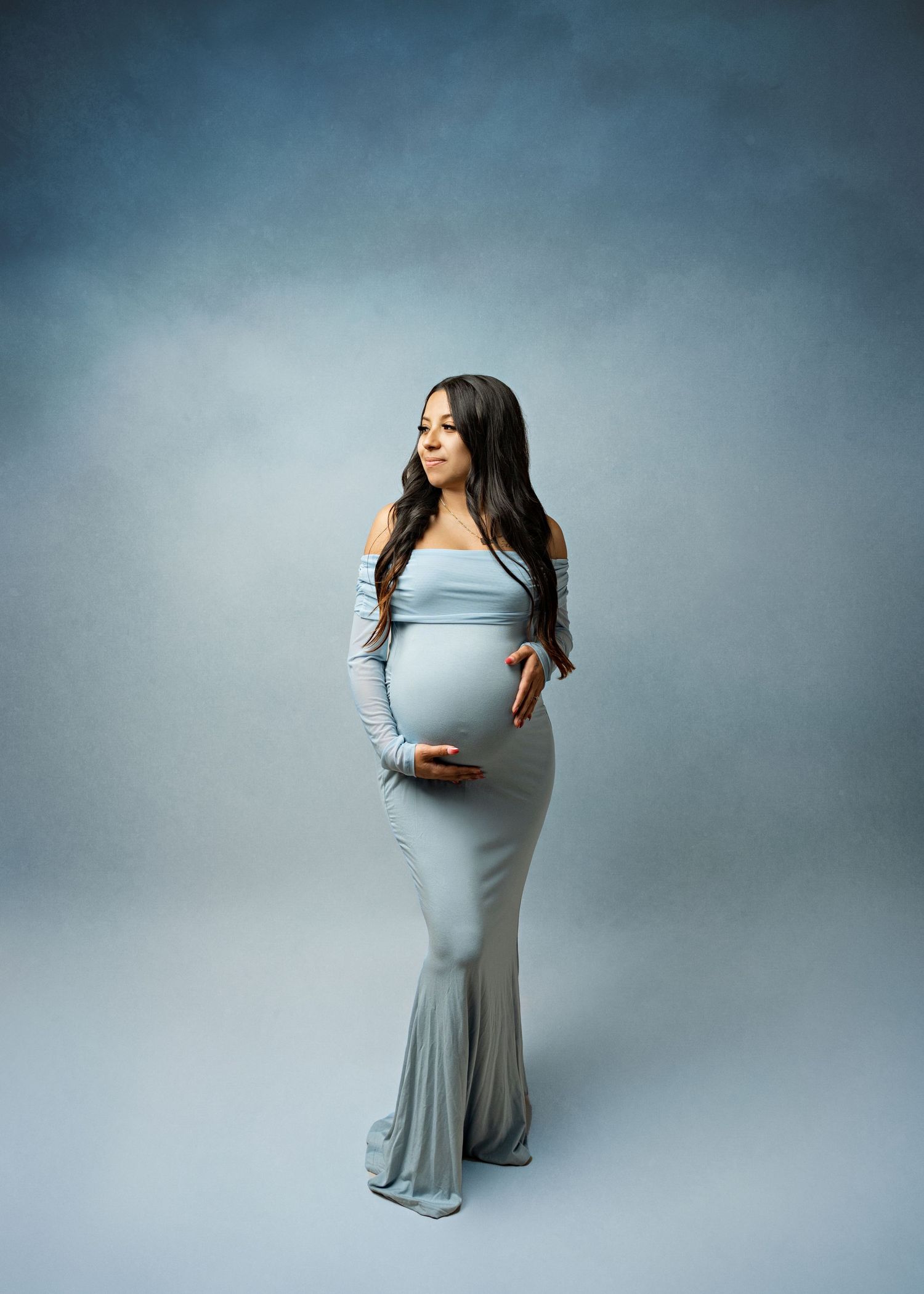 Elegant maternity portrait of person wearing a flowing light blue floor-length gown in a moody studio setting.