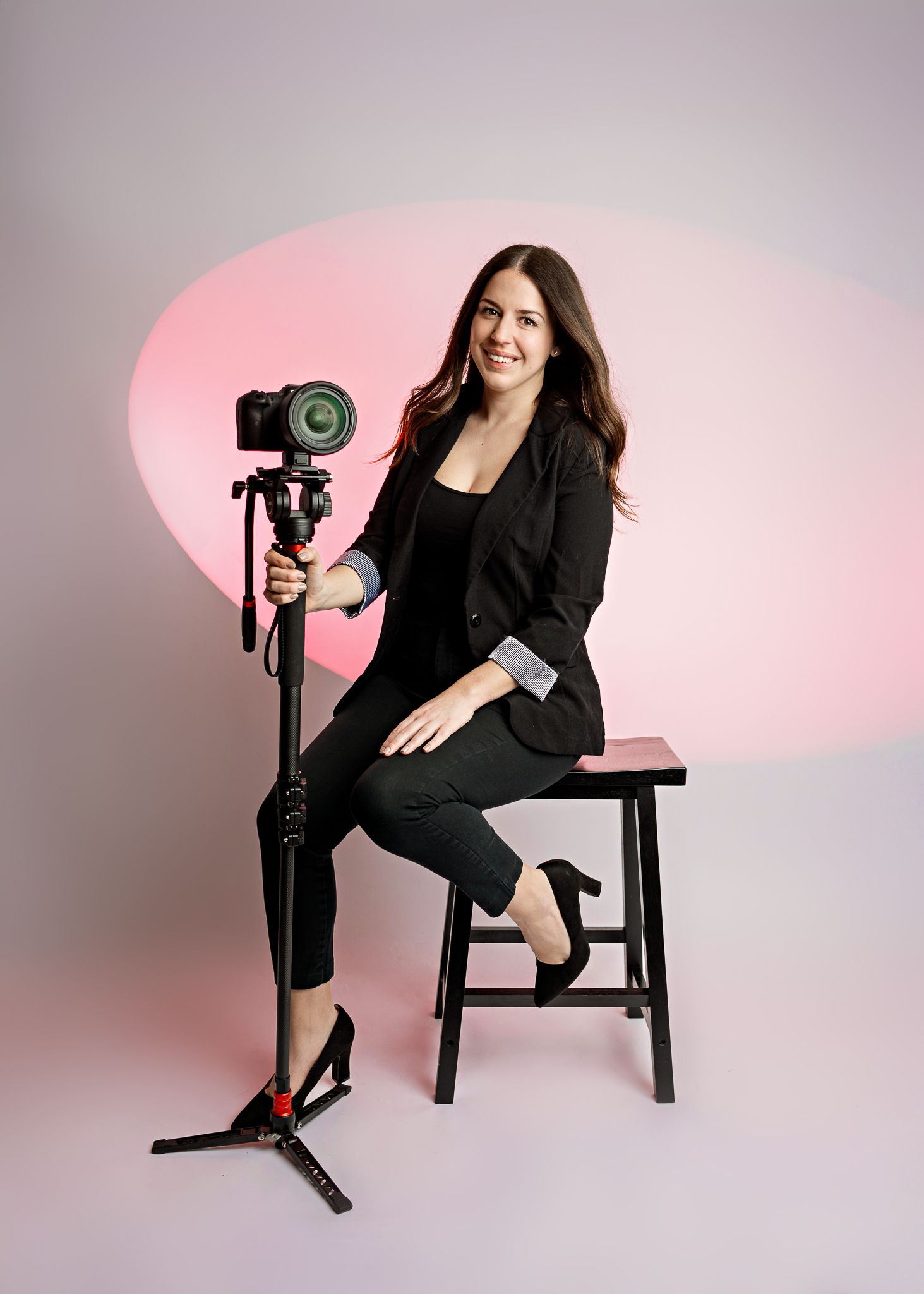 Professional photographer in all black business attire poses on stool with camera mounted on tripod against pink background.