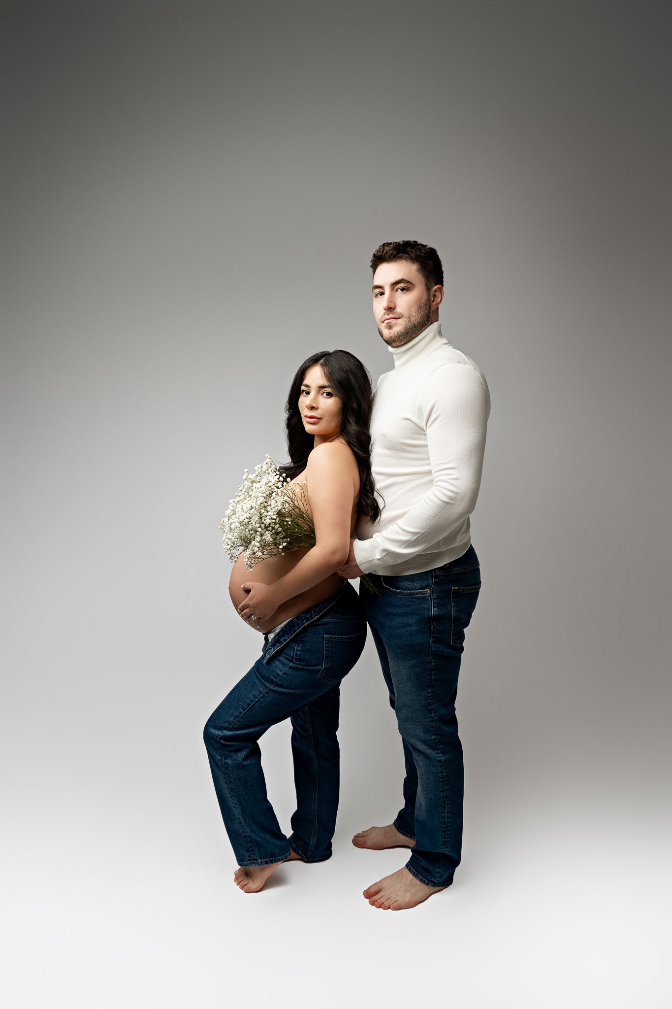 A couple in white and jeans pose back to back in a studio setting with baby's breath flowers.