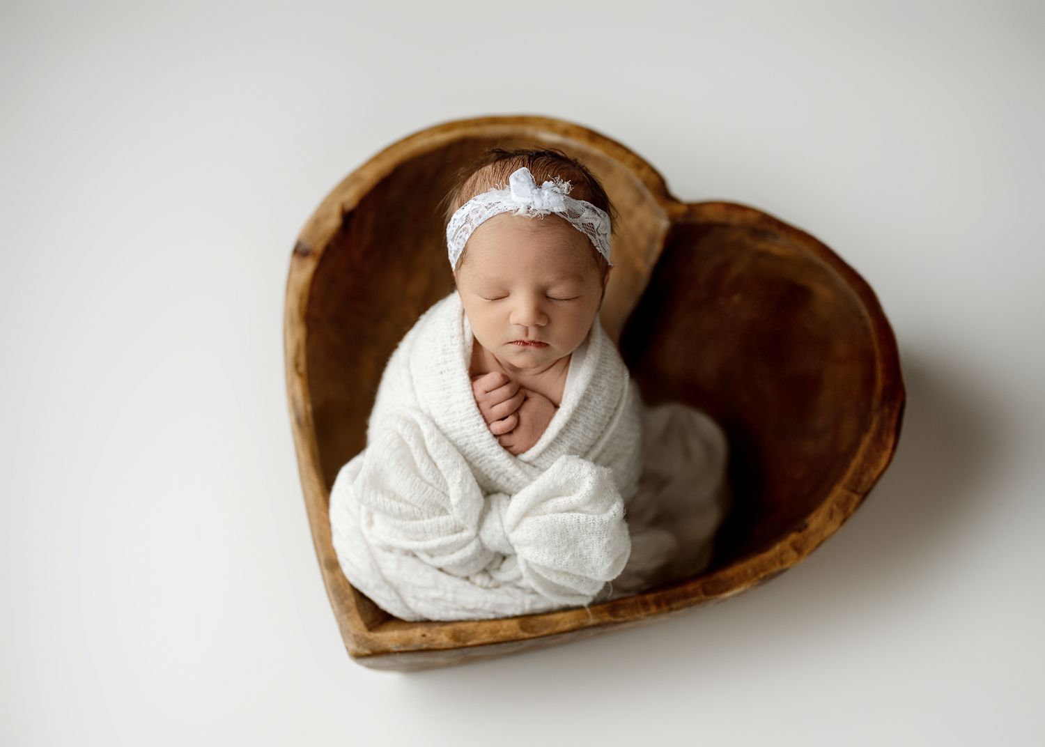 A series of five photos showing a sleeping newborn baby wrapped in white cloth and nestled in a wooden heart-shaped bowl.