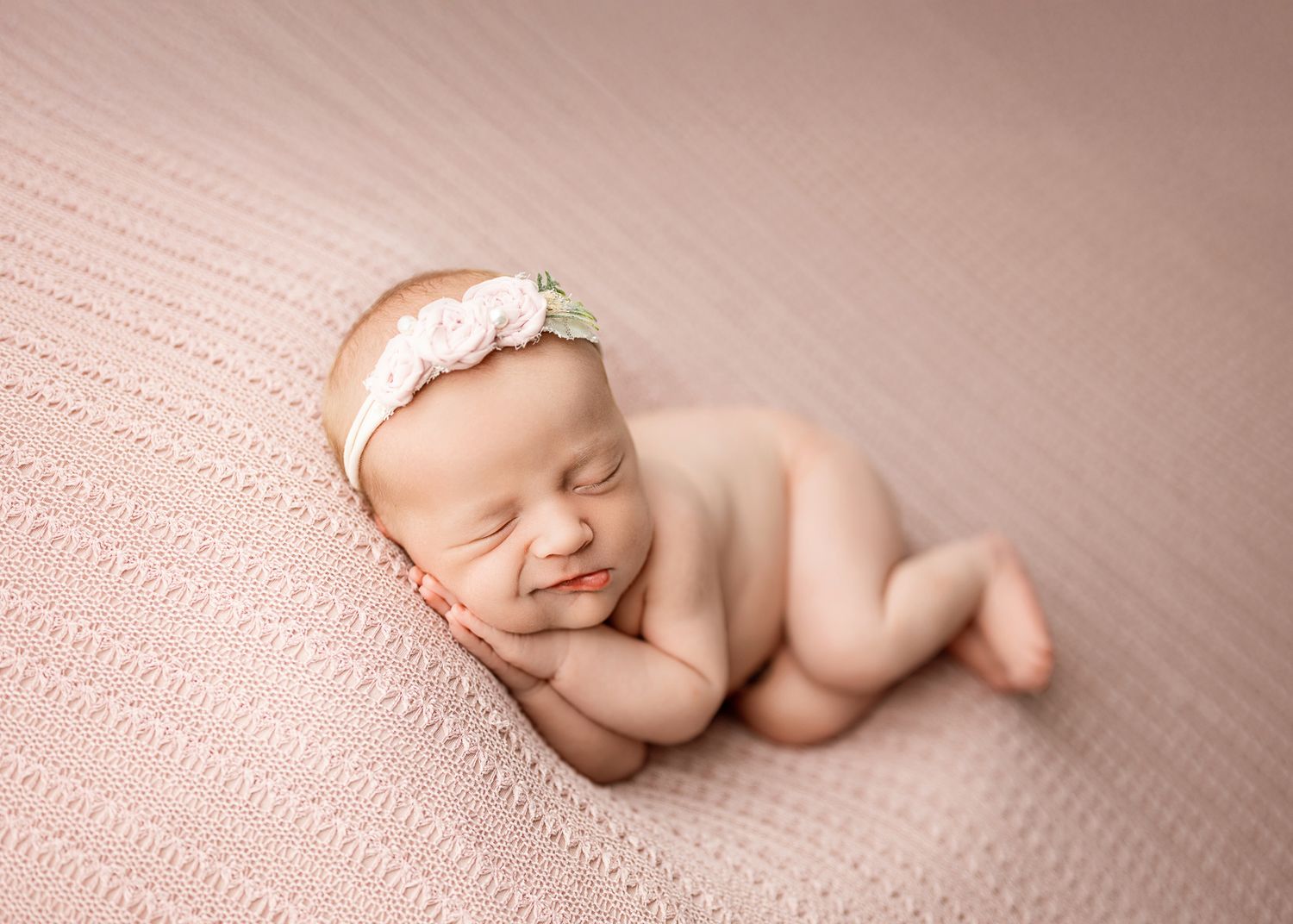 Peaceful sleeping newborn baby wearing delicate lace headband on soft pink textured blanket.