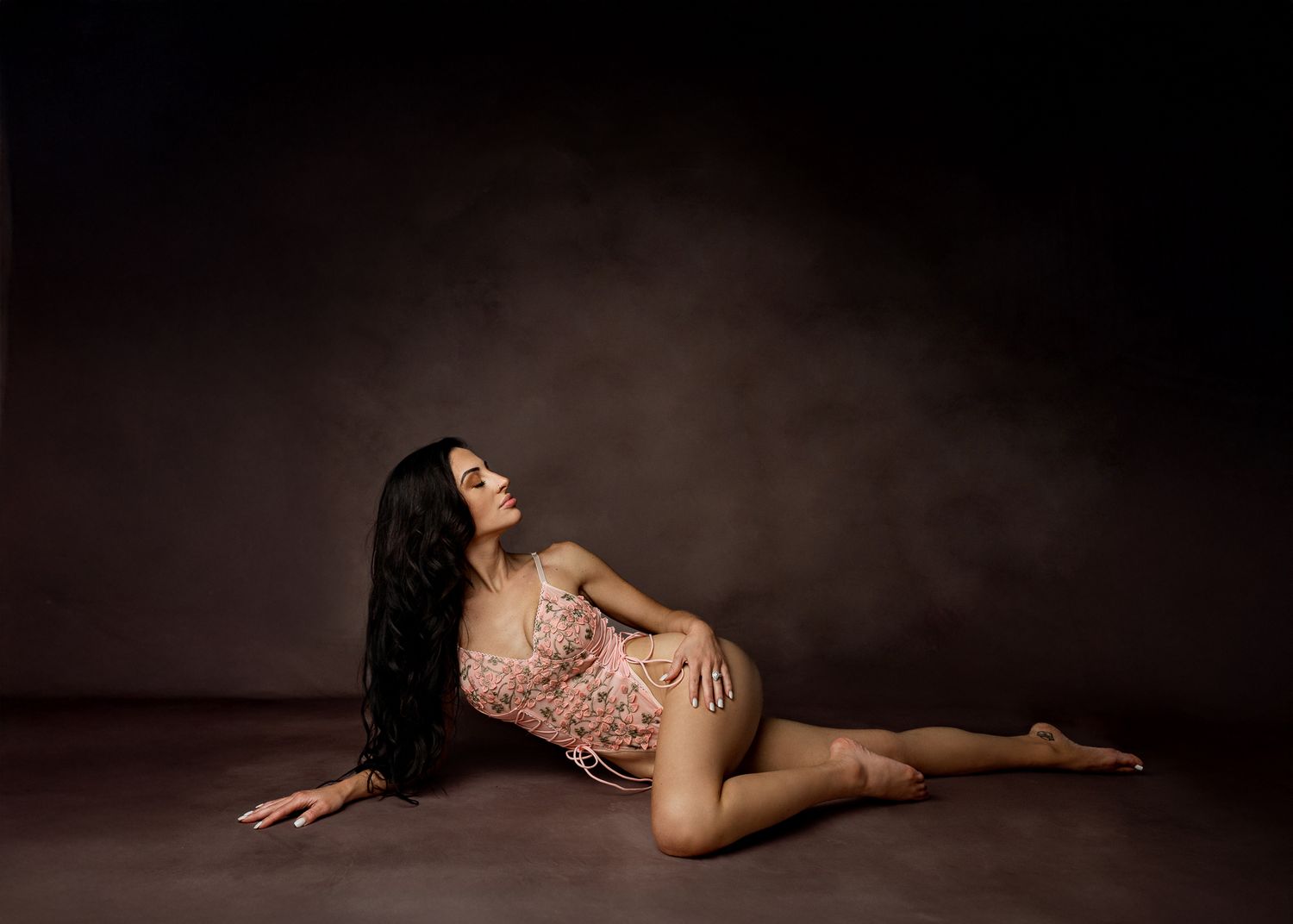 Artistic maternity photo of woman in pink lace dress reclining on dark backdrop in professional studio setting.
