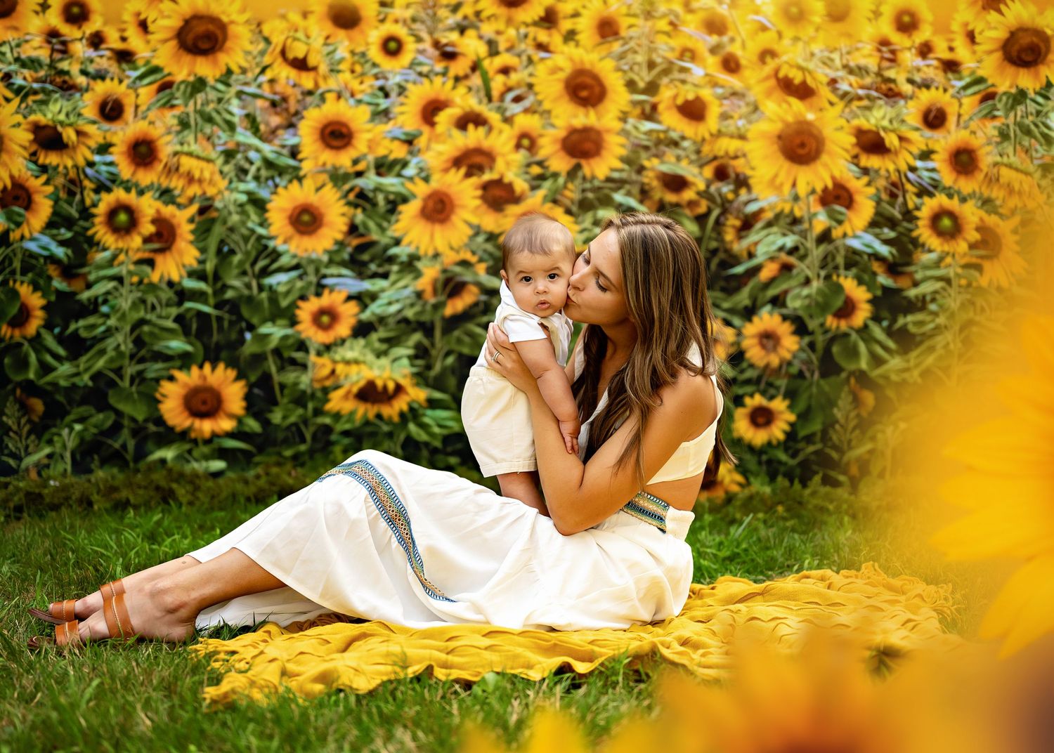 Two people sitting together on a blanket surrounded by vibrant yellow sunflowers in a garden setting.