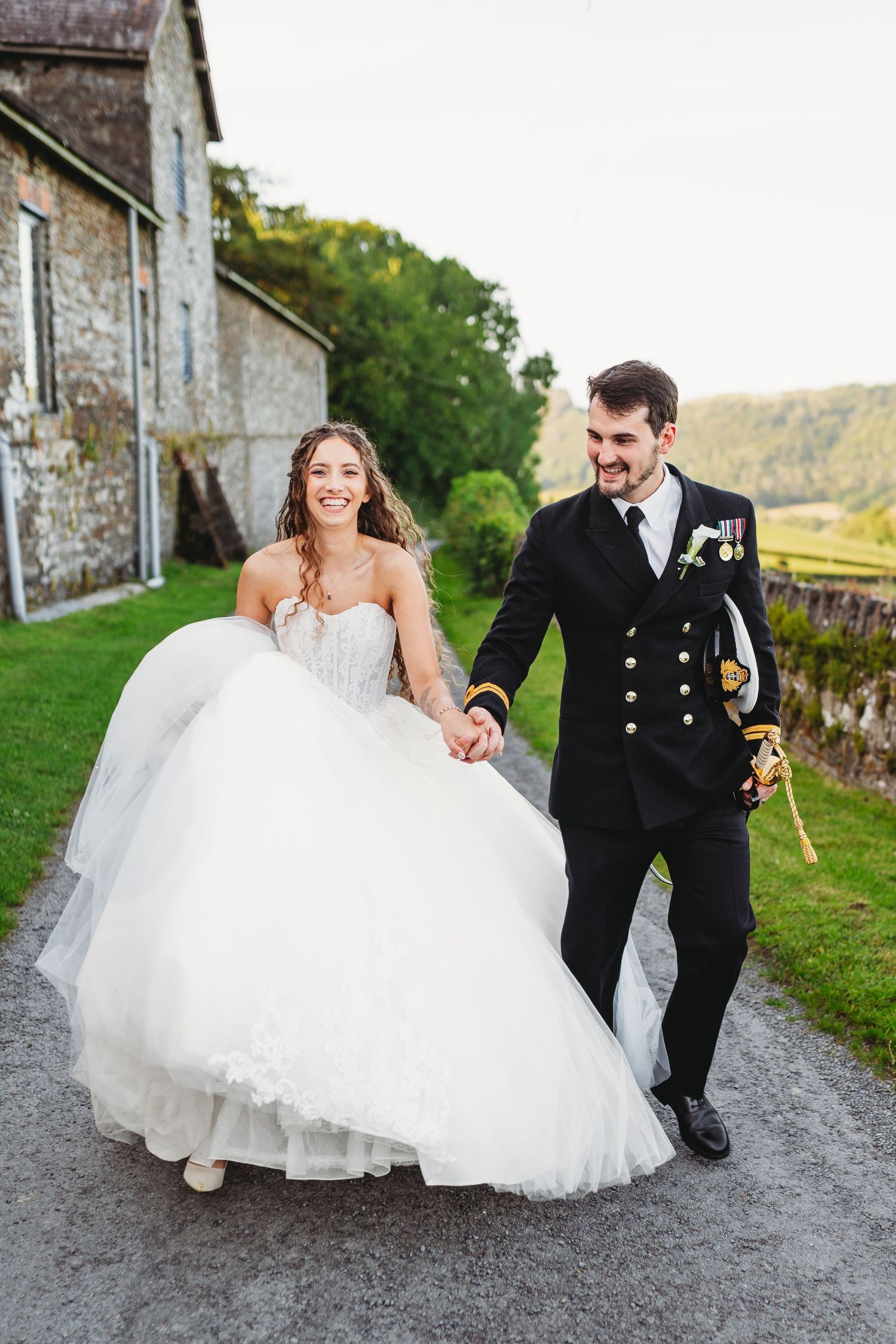 Smiling bride in a white tulle ball gown holds hands with a groom in a navy military dress uniform while walking along a country path.