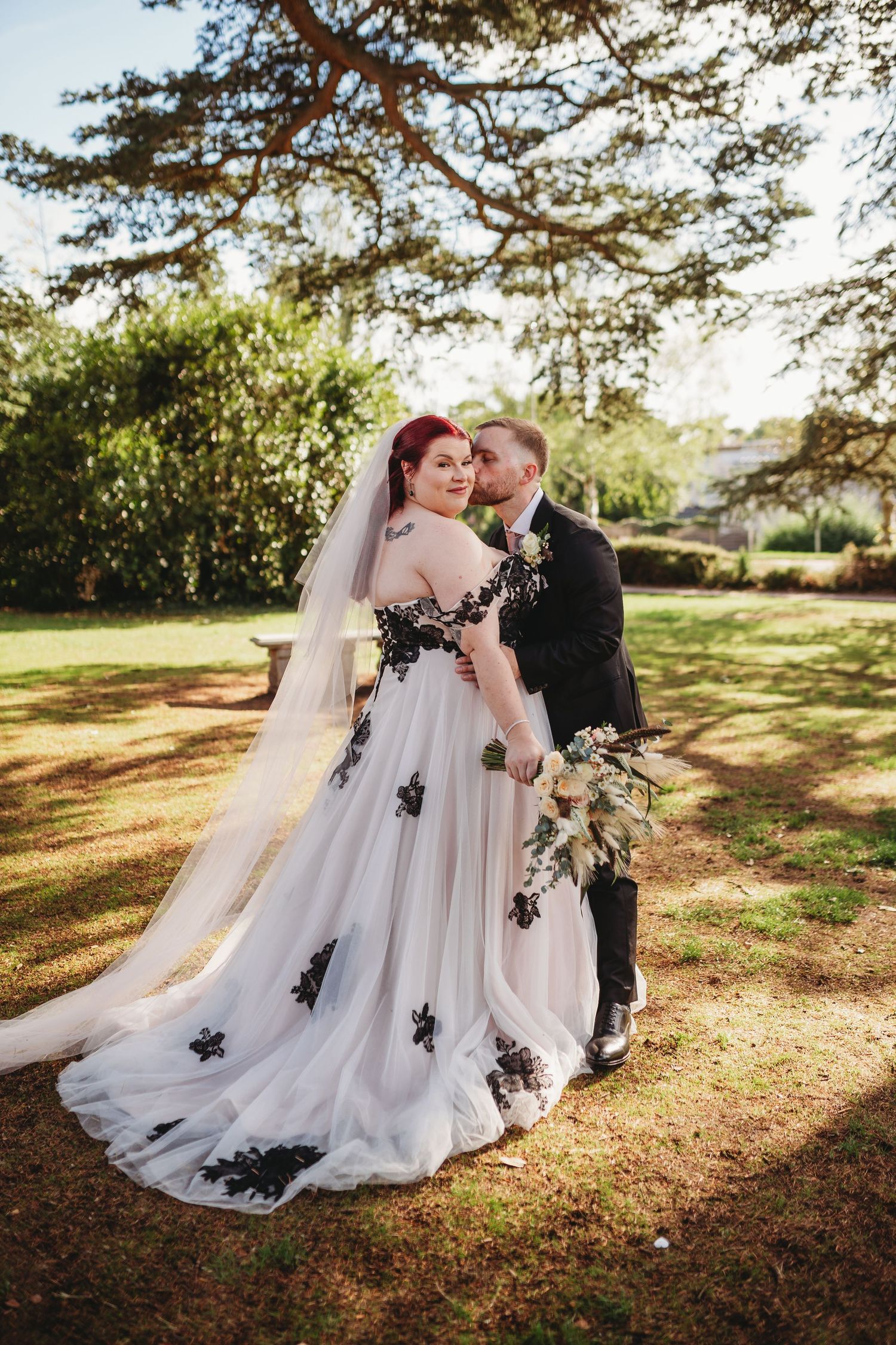 Bride in a white tulle gown with black floral appliqués and long veil holds a bouquet while the groom kisses her cheek in a sunny park.