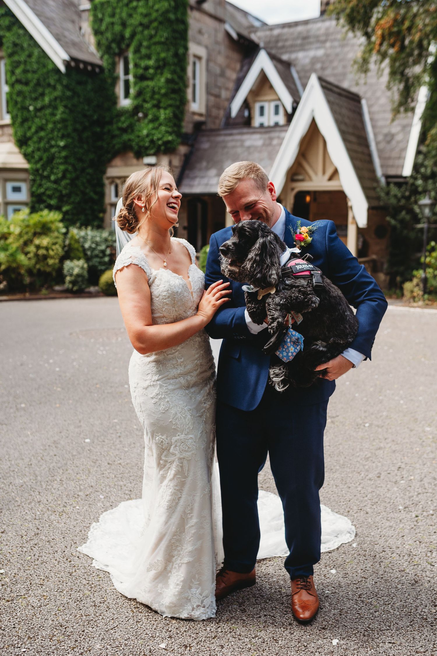 A laughing bride in a lace gown and a groom in a navy suit hold their fluffy black dog outside an ivy-covered stone wedding venue.