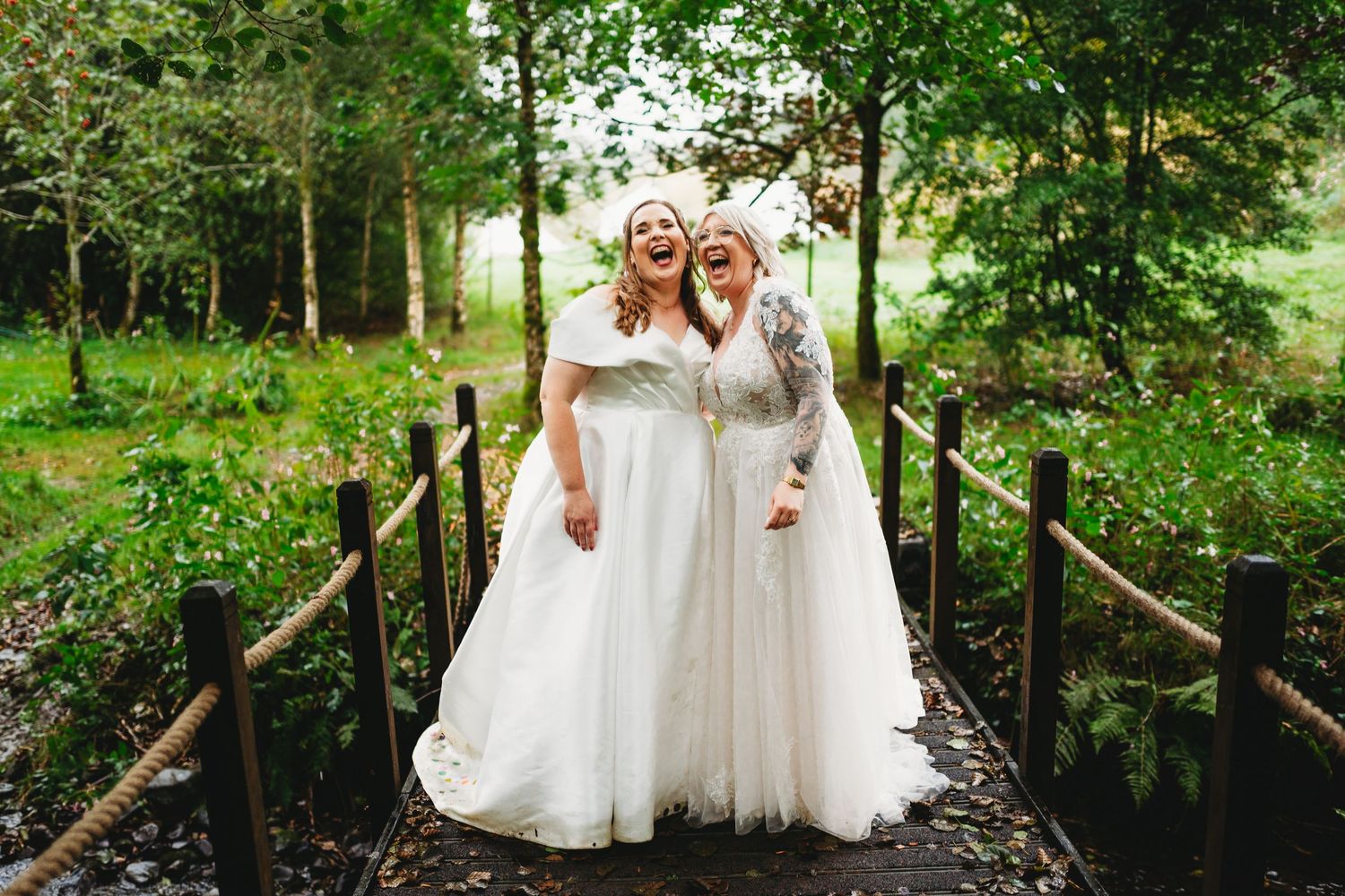 Two brides in white wedding gowns laugh together on a wooden footbridge with rope railings, surrounded by lush green woodland.