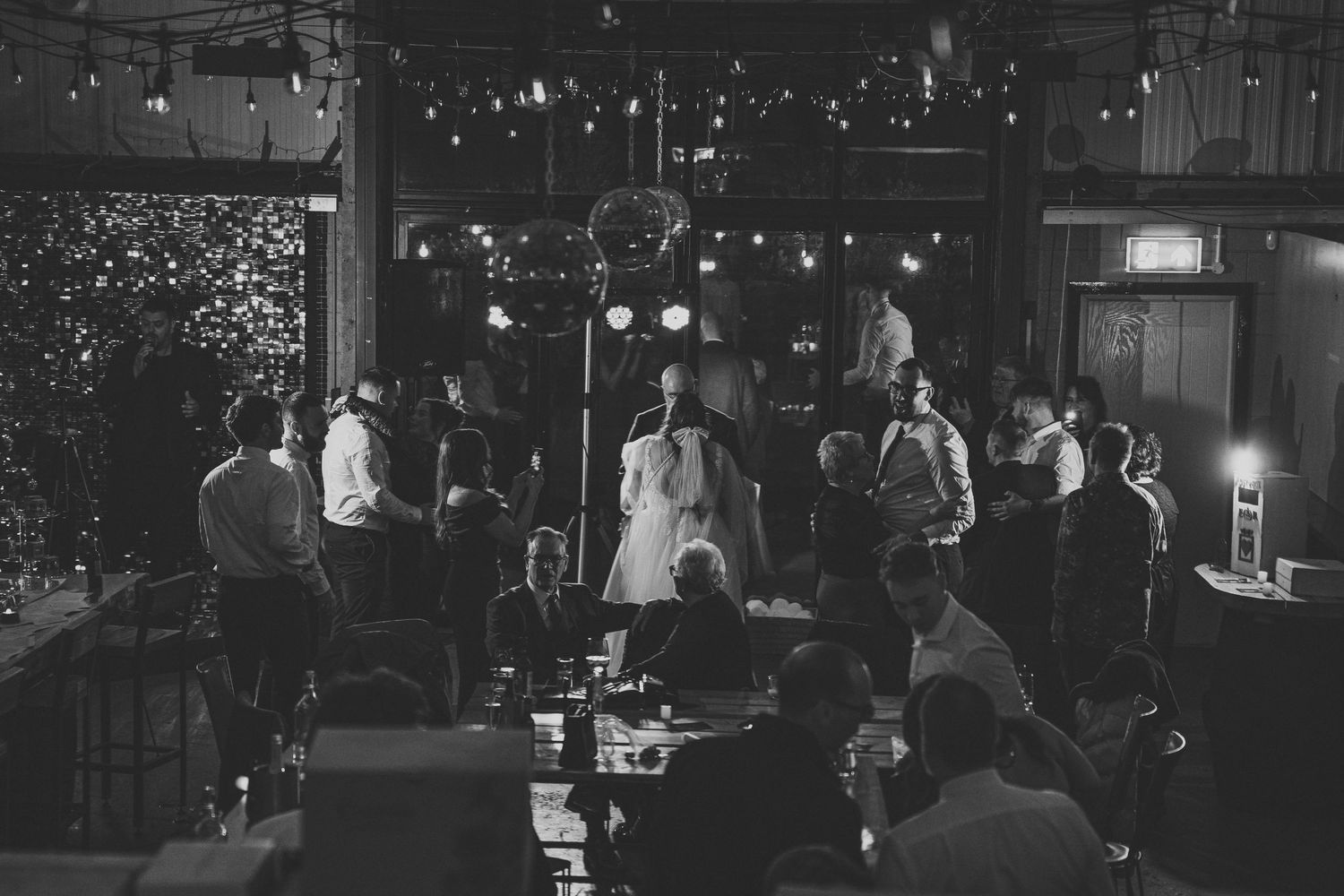 Black and white photo of wedding guests gathered around tables as a couple shares a first dance in a softly lit venue with string lights.