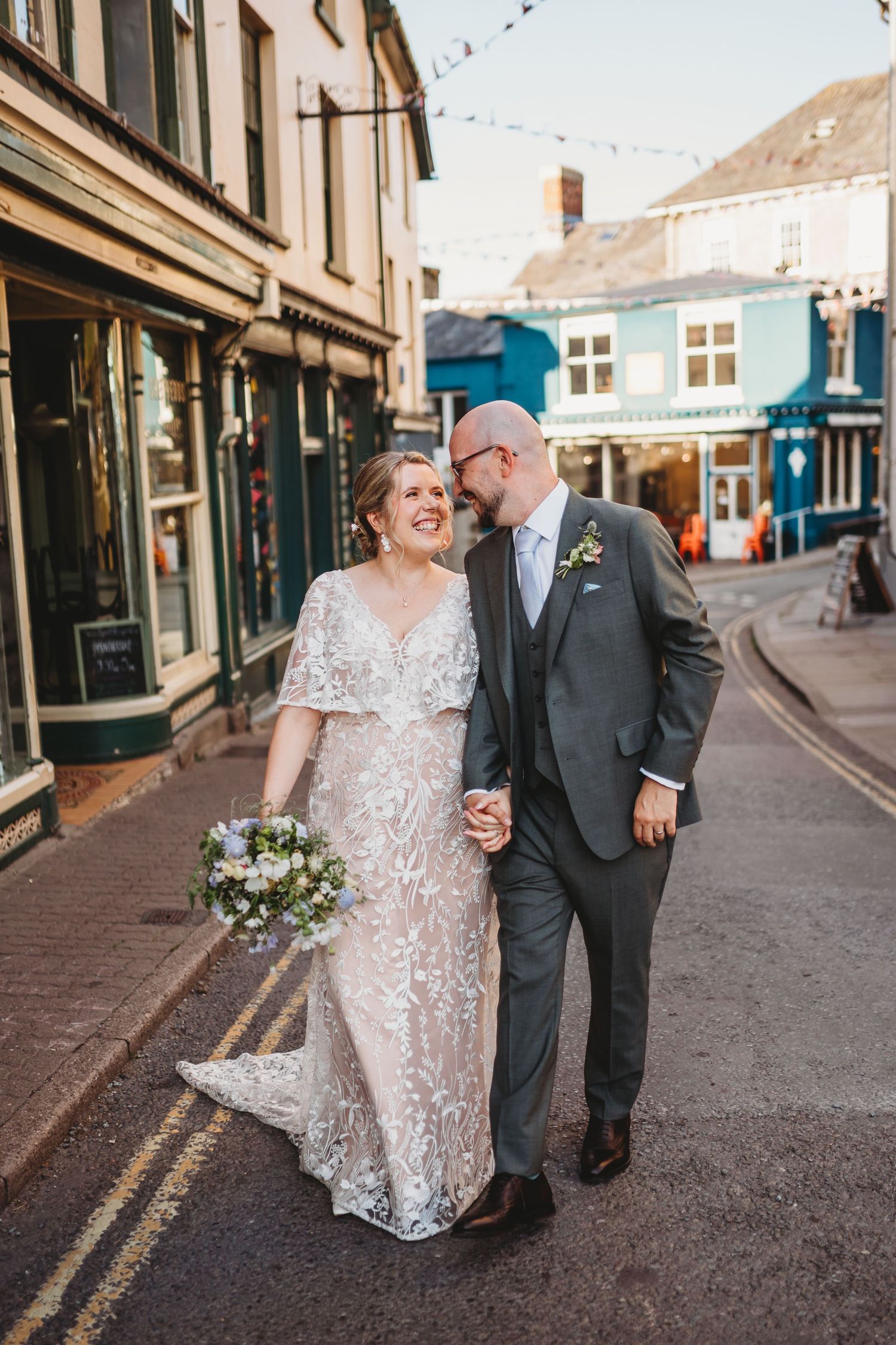 Smiling bride in a lace wedding gown and groom in a grey suit walk hand in hand down a charming street, holding a bouquet.