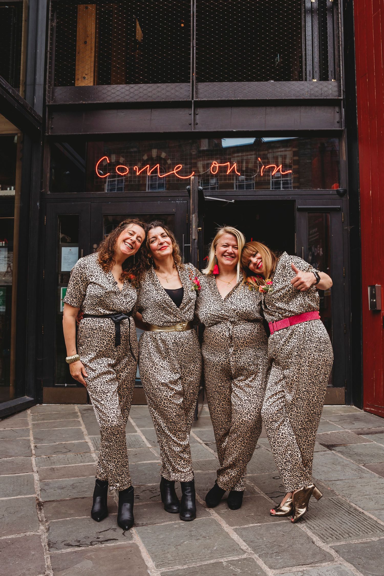 Four women in matching leopard print jumpsuits pose together outside a venue with a neon 'Come On In' sign.