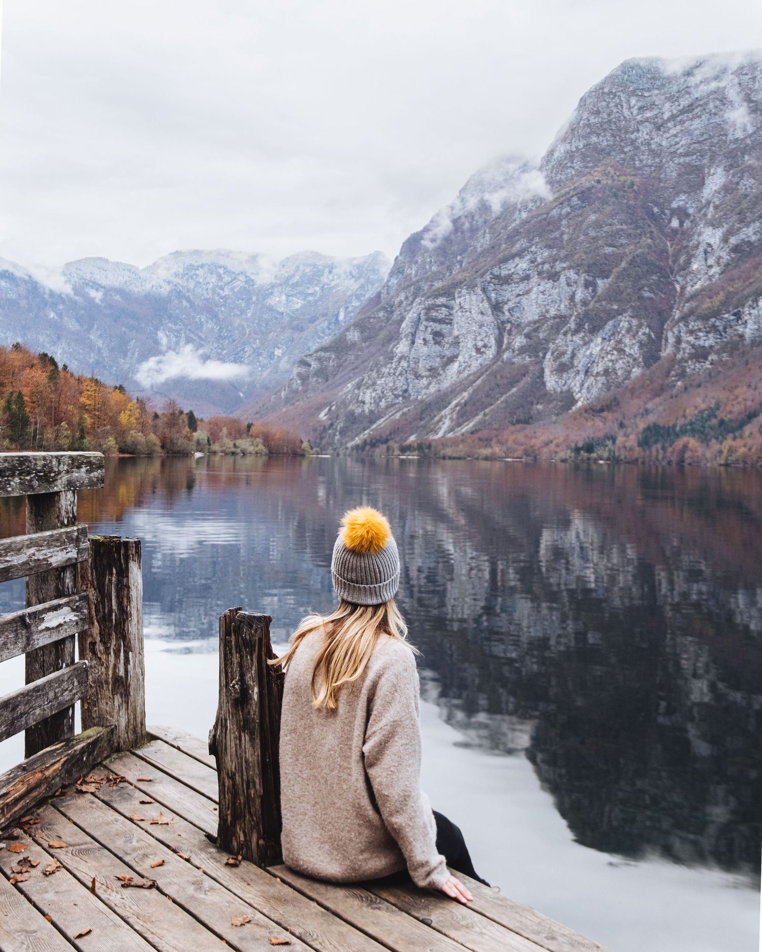 A girl sits on a dock looking out over a lake and mountains.