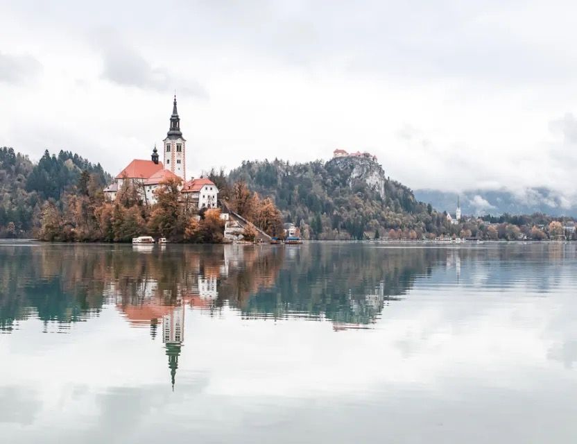 Snowy lakeside church reflected in calm water with mountains behind.