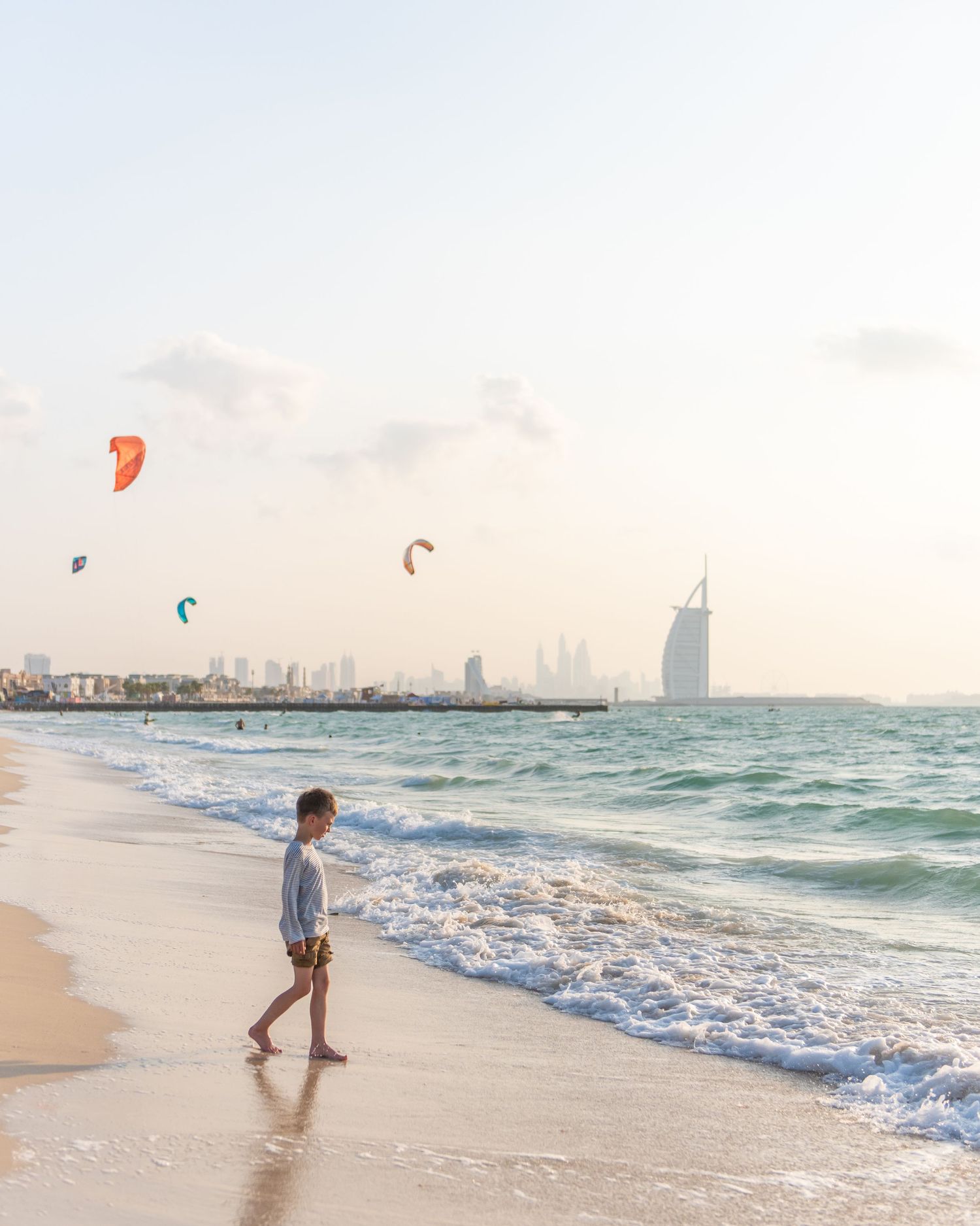 Person walking along Dubai beach with kitesurfers and Burj Al Arab hotel visible in the distance during sunset.
