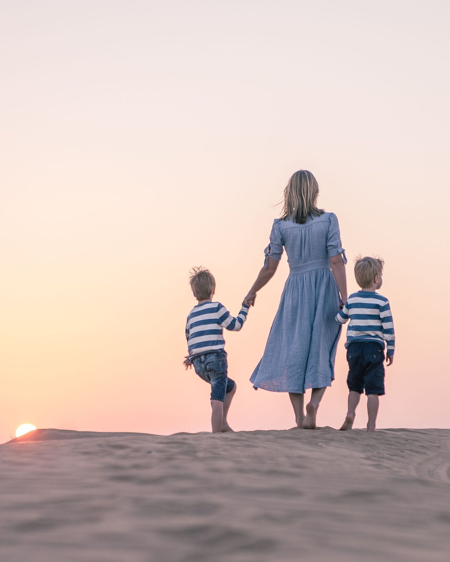 A figure in a blue dress holds hands with two small children in striped shirts watching sunset on sand dunes.