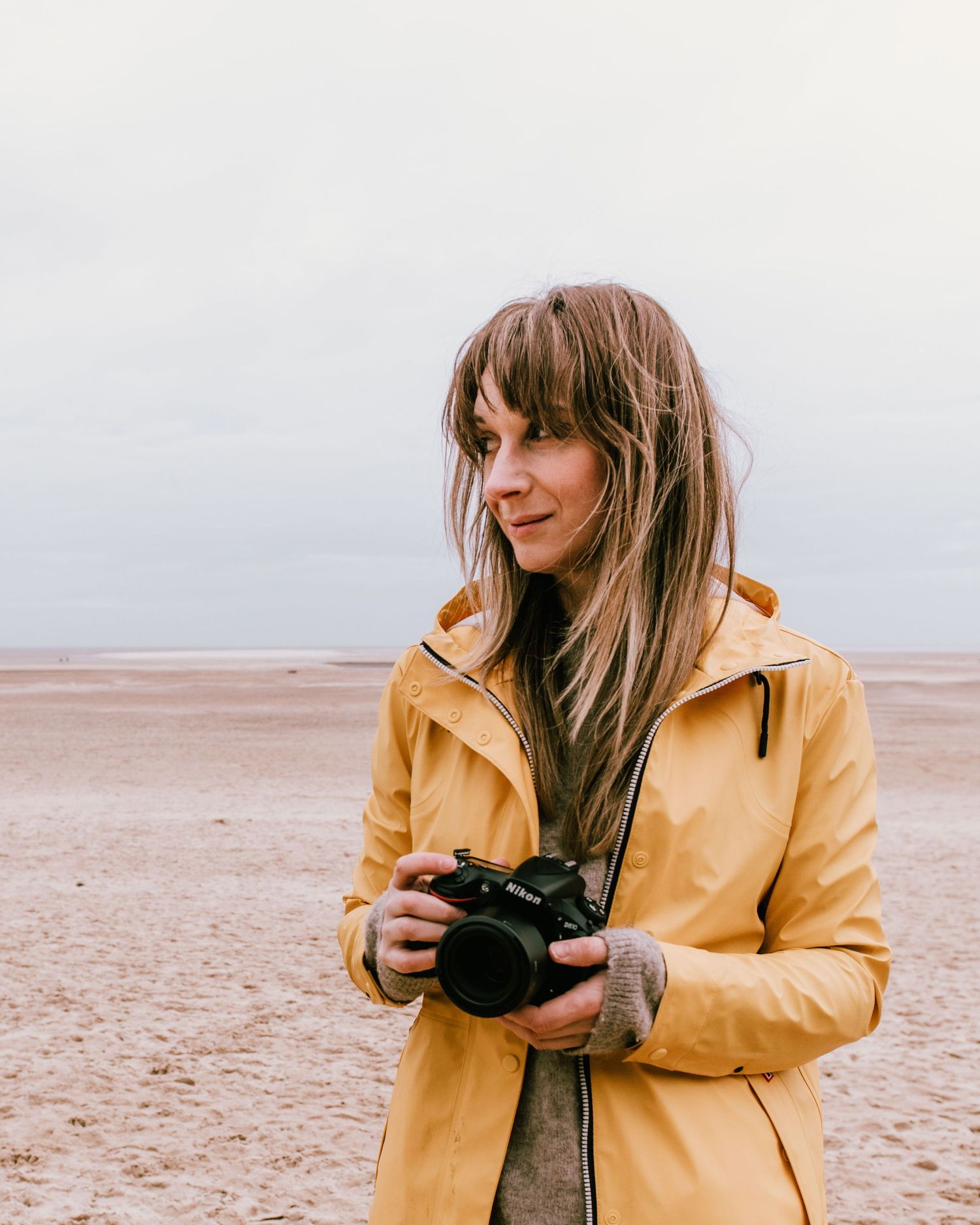A photographer wearing a yellow raincoat holds a vintage camera while exploring a misty beach landscape.