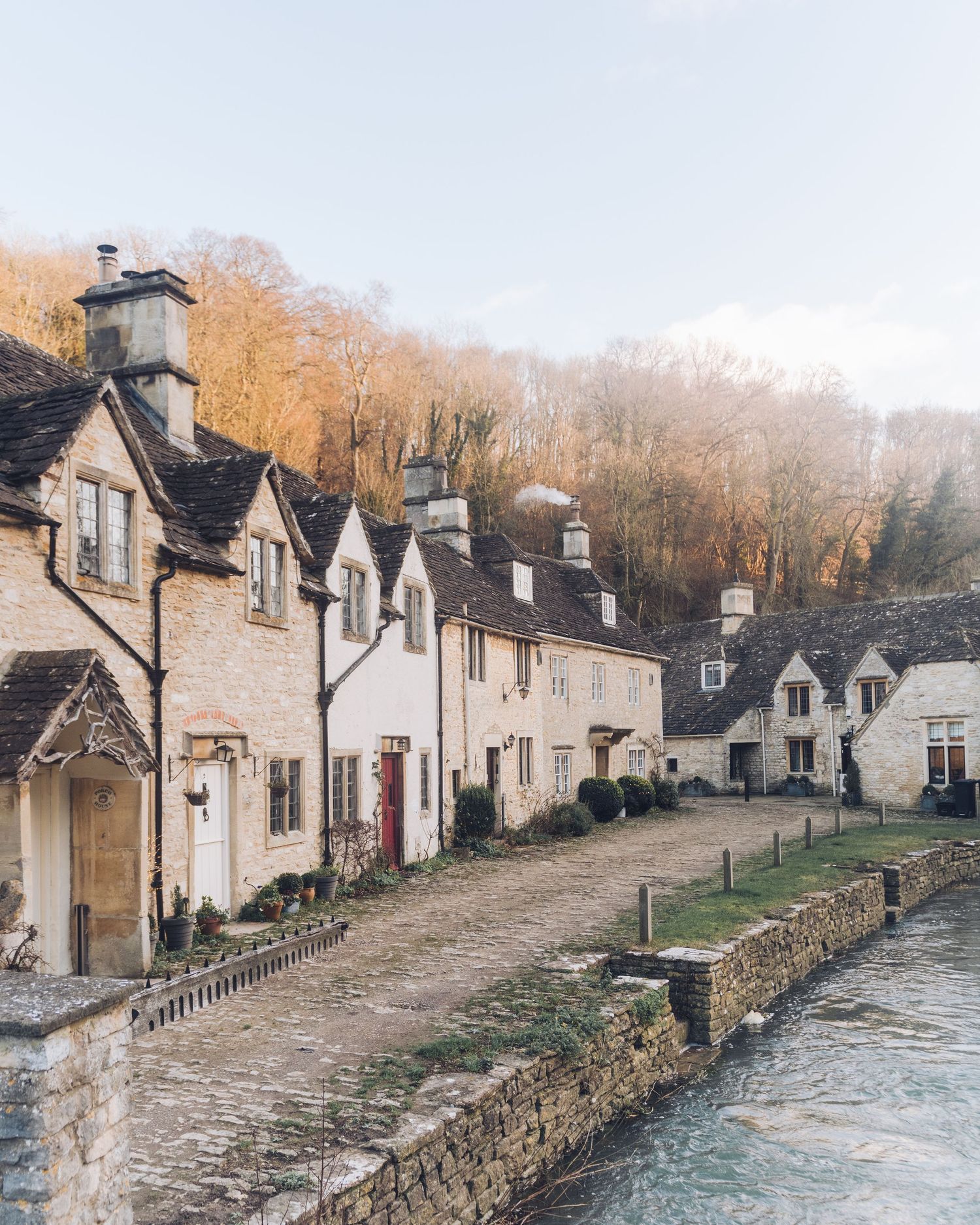 Charming stone cottages line a rustic street along a river in a quaint European village during autumn.