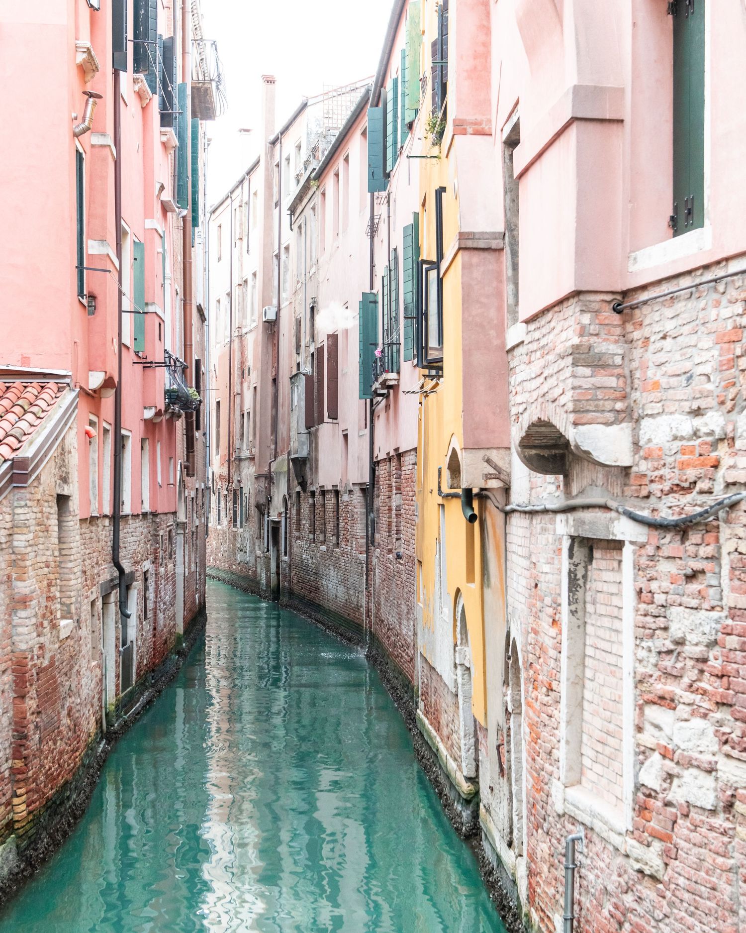 Narrow Venetian canal with turquoise water flowing between weathered pink and yellow buildings with exposed brick walls.