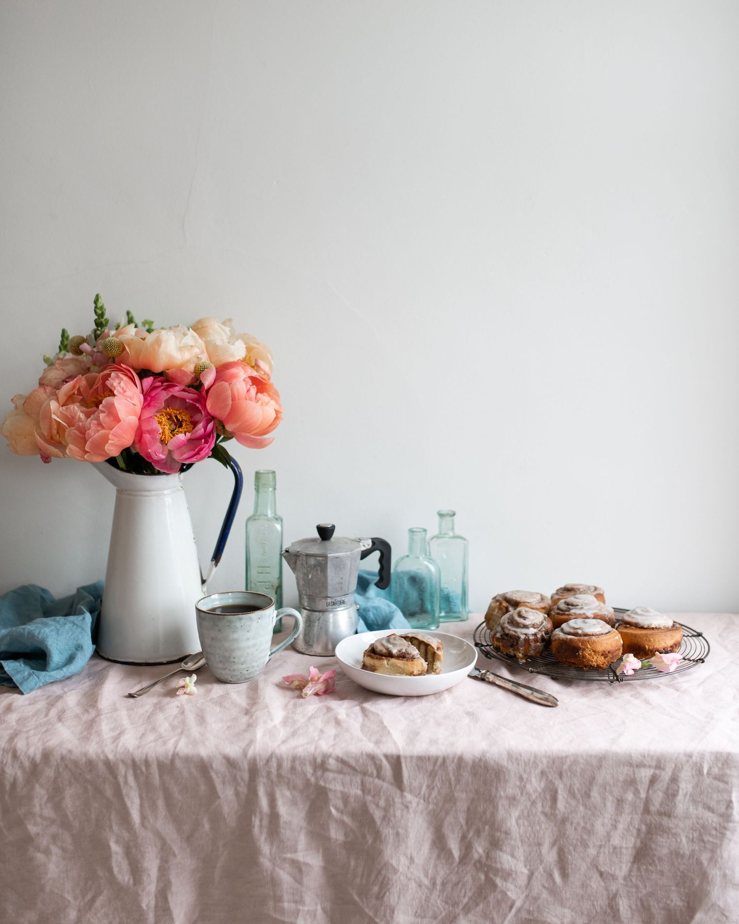 A rustic table setting with pink flowers in vases, pastries, and glassware arranged on a light linen tablecloth.