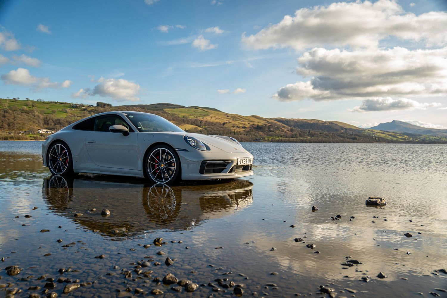 Sleek silver Porsche sports car parked on wet beach with mountains and reflective water in background.